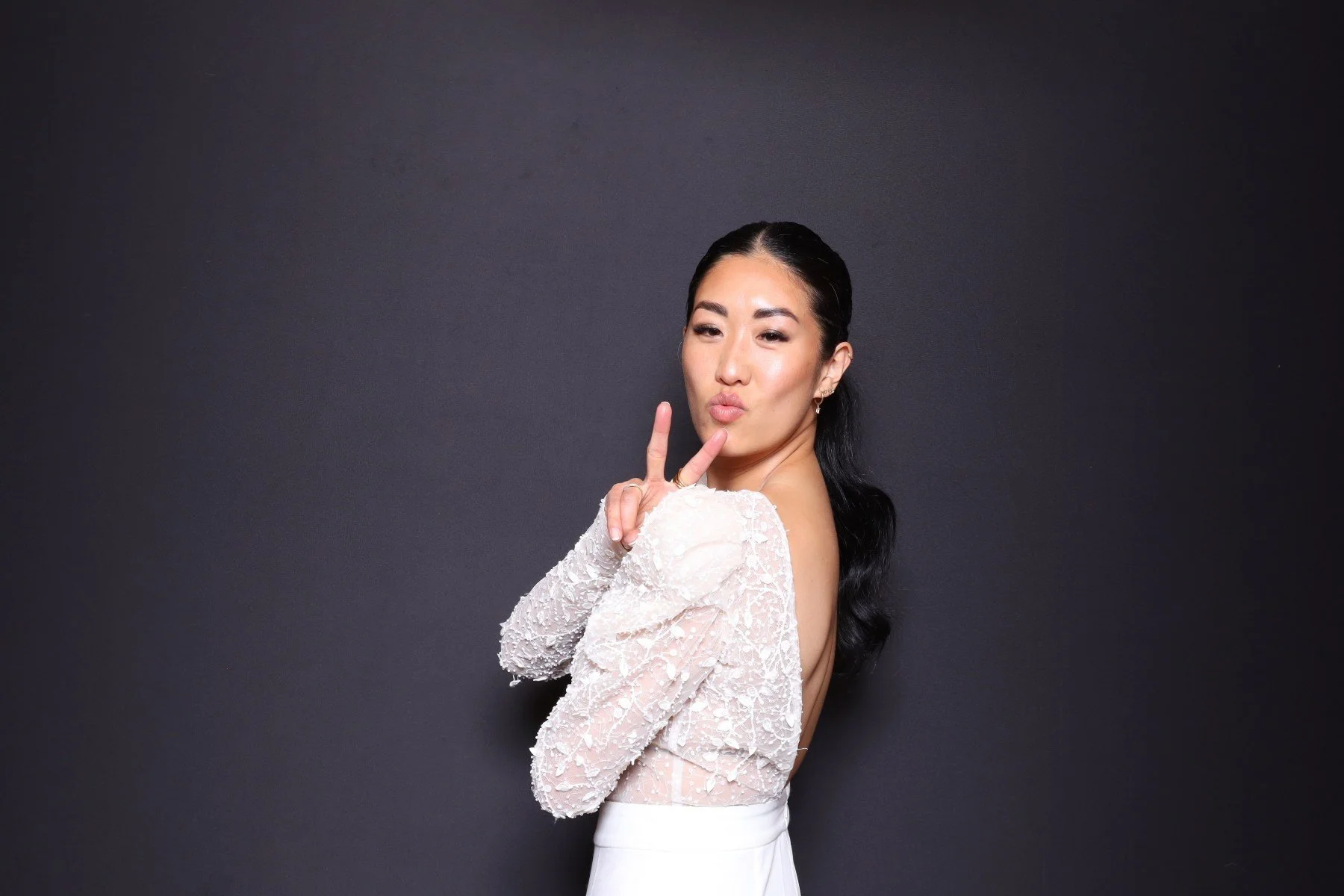 Portrait of a young Asian woman with long black hair, wearing a white lace dress, making a peace sign with her right hand and pouting her lips against a dark background.