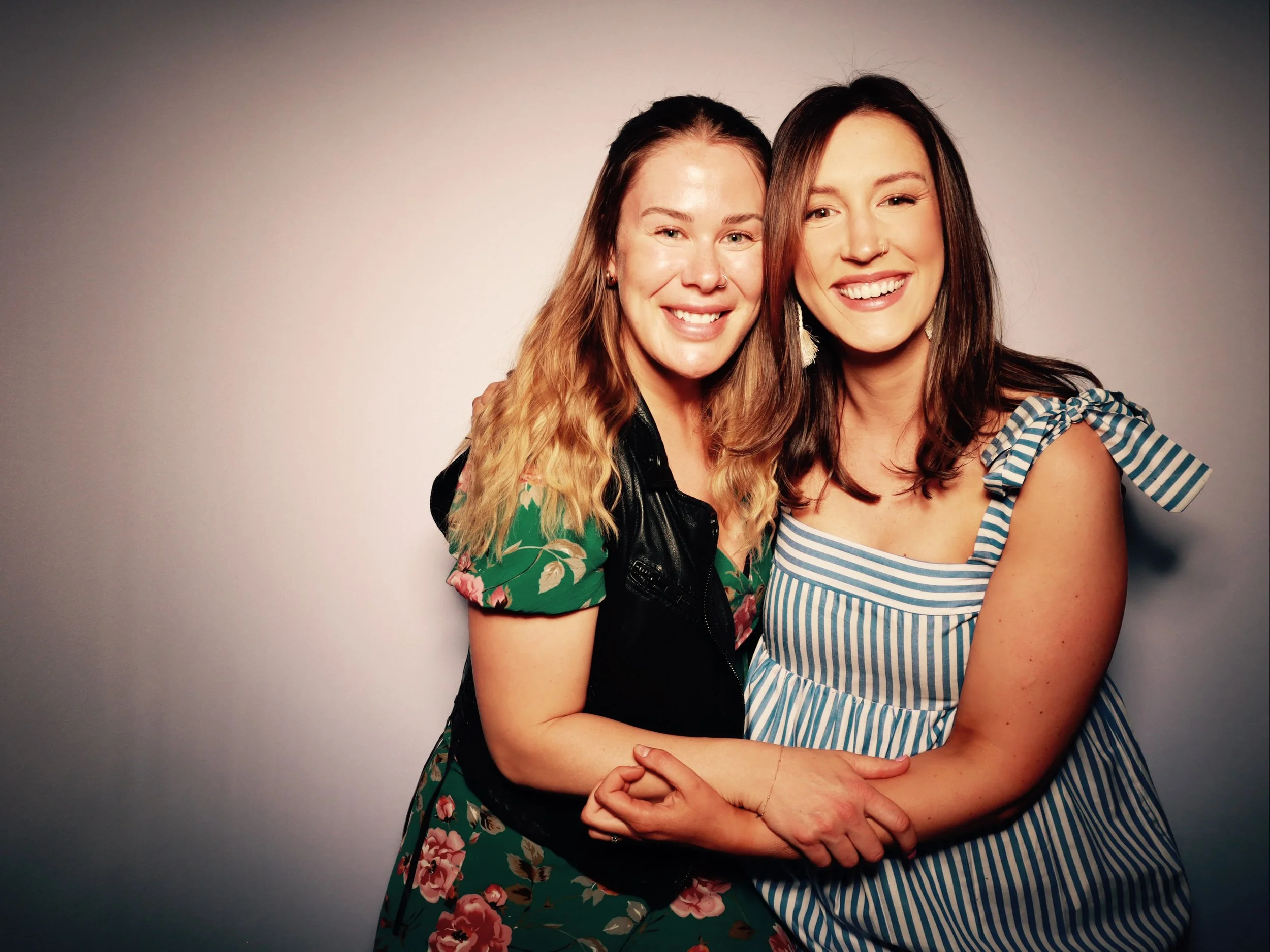 Two women smiling and embracing each other in front of a plain background.