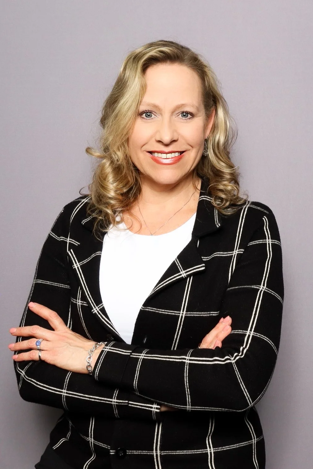 A smiling blonde woman with wavy hair wearing a black blazer with white plaid pattern, crossed arms, and a white shirt, standing against a plain light purple background.