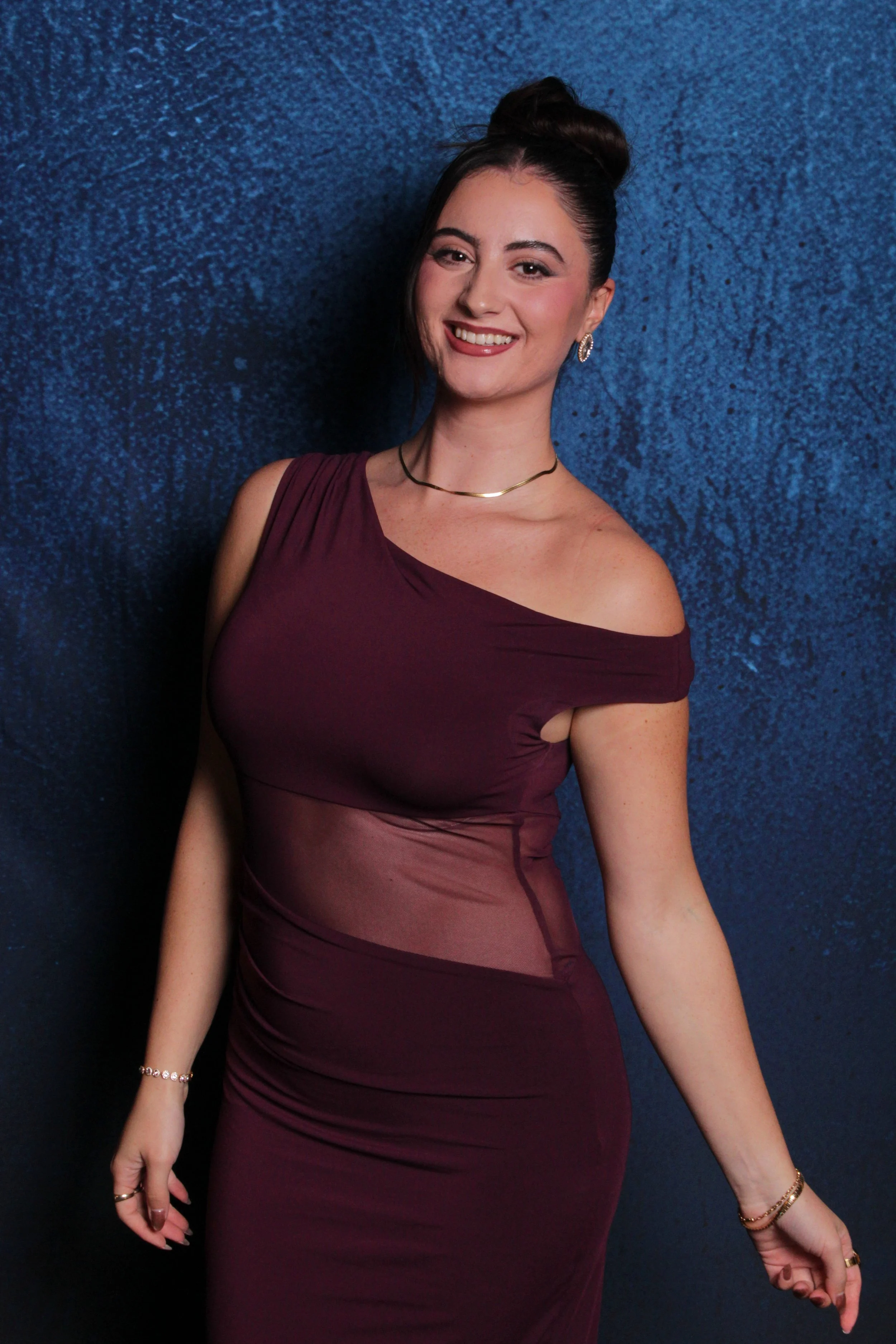 A woman smiling and posing against a textured blue background, wearing a burgundy dress with a sheer panel at the waist, jewelry, and her hair styled in an updo.