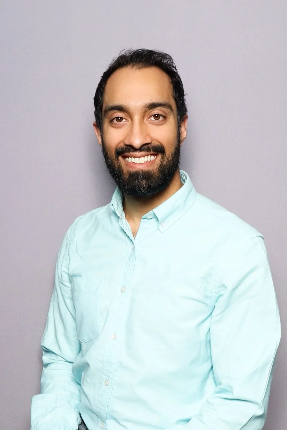 A man with dark hair, beard, and mustache smiling, wearing a light blue button-up shirt, standing against a plain light gray background.