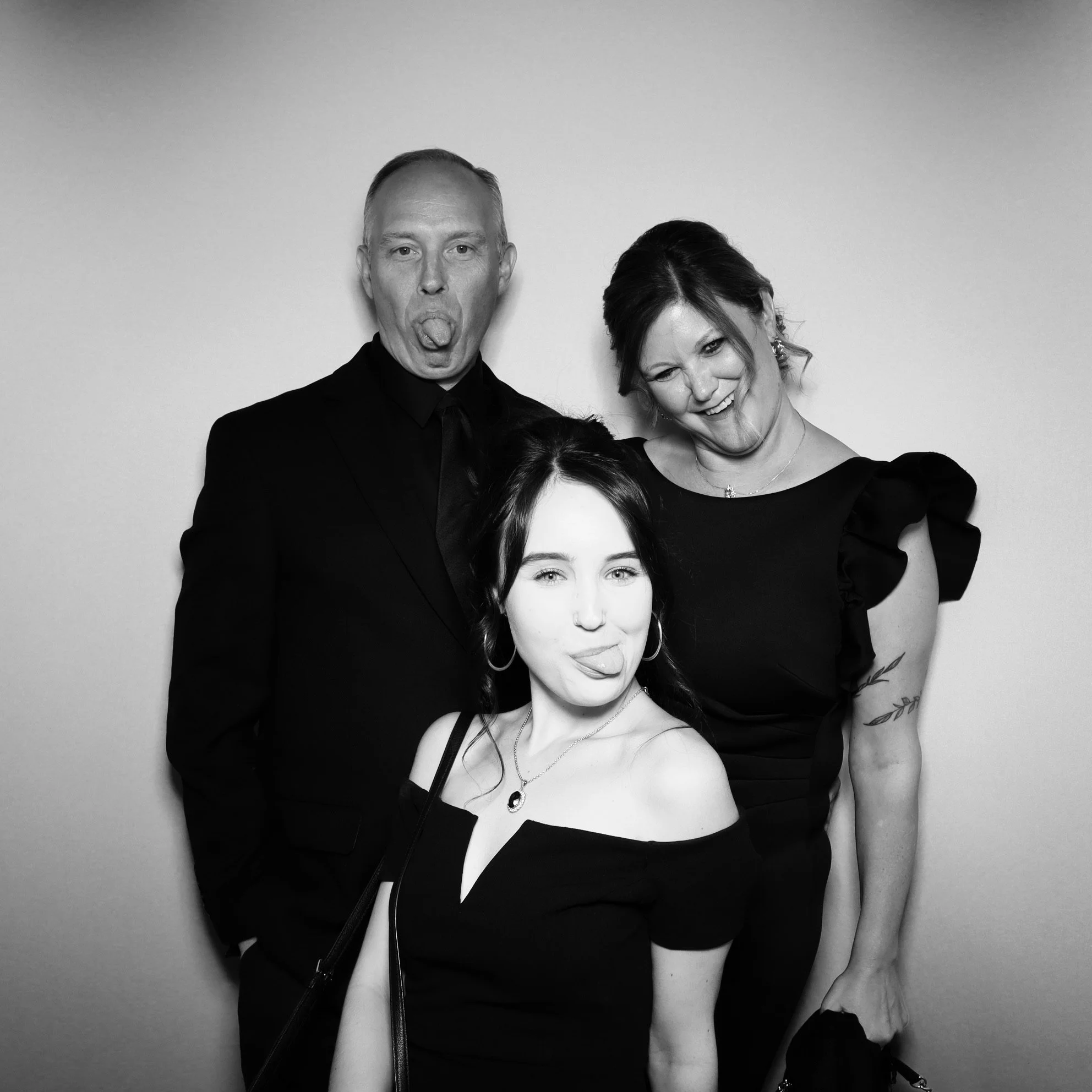 Three people in black attire making playful faces in a photo booth, with a plain background.