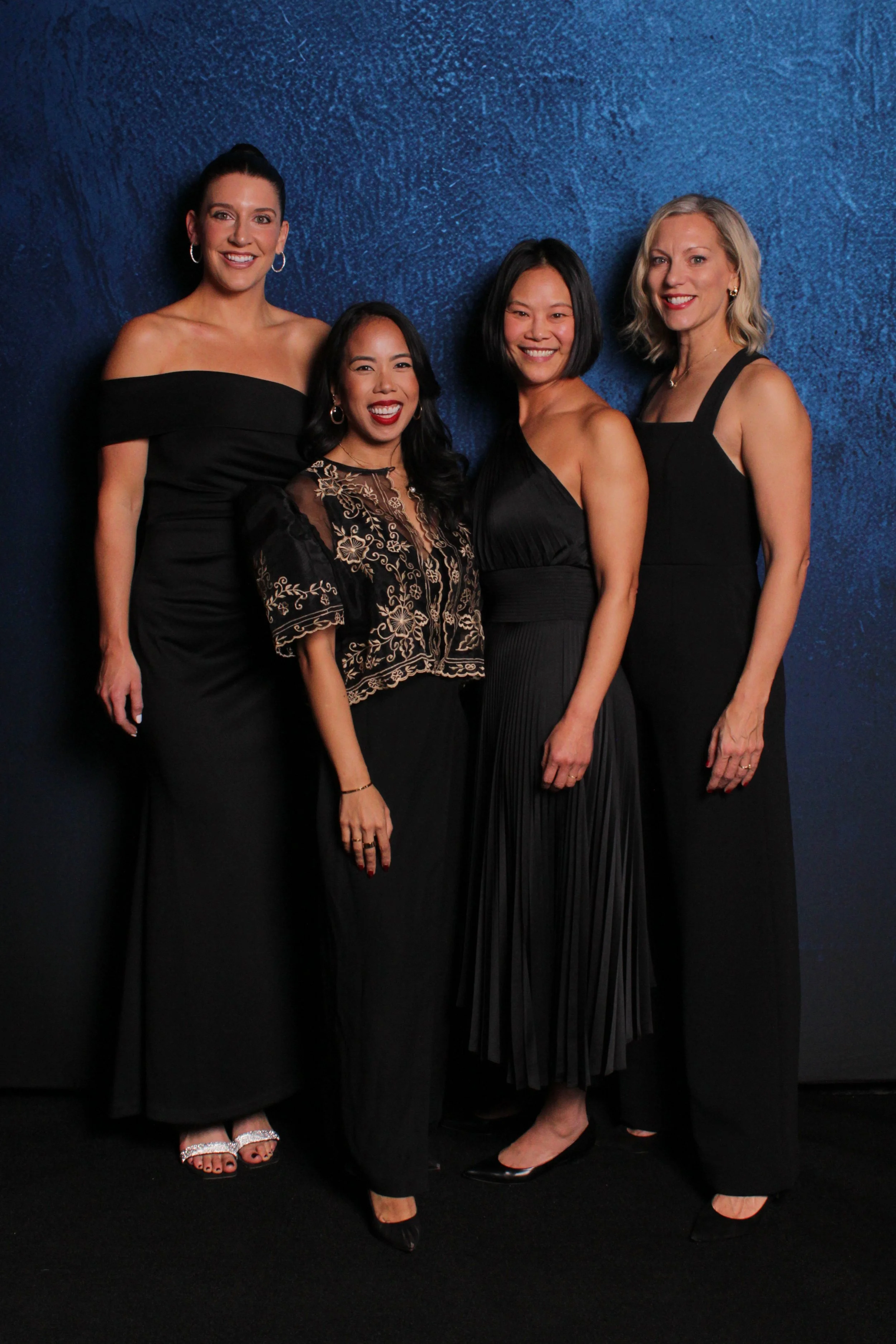 A group of four women dressed in elegant black attire, standing against a blue textured backdrop.