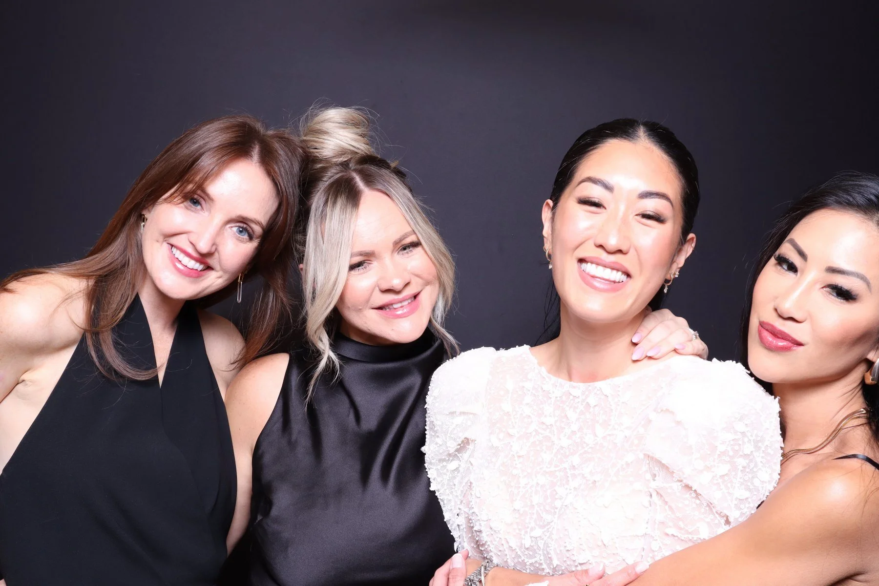 Four women smiling and posing together against a dark background, dressed in elegant attire.
