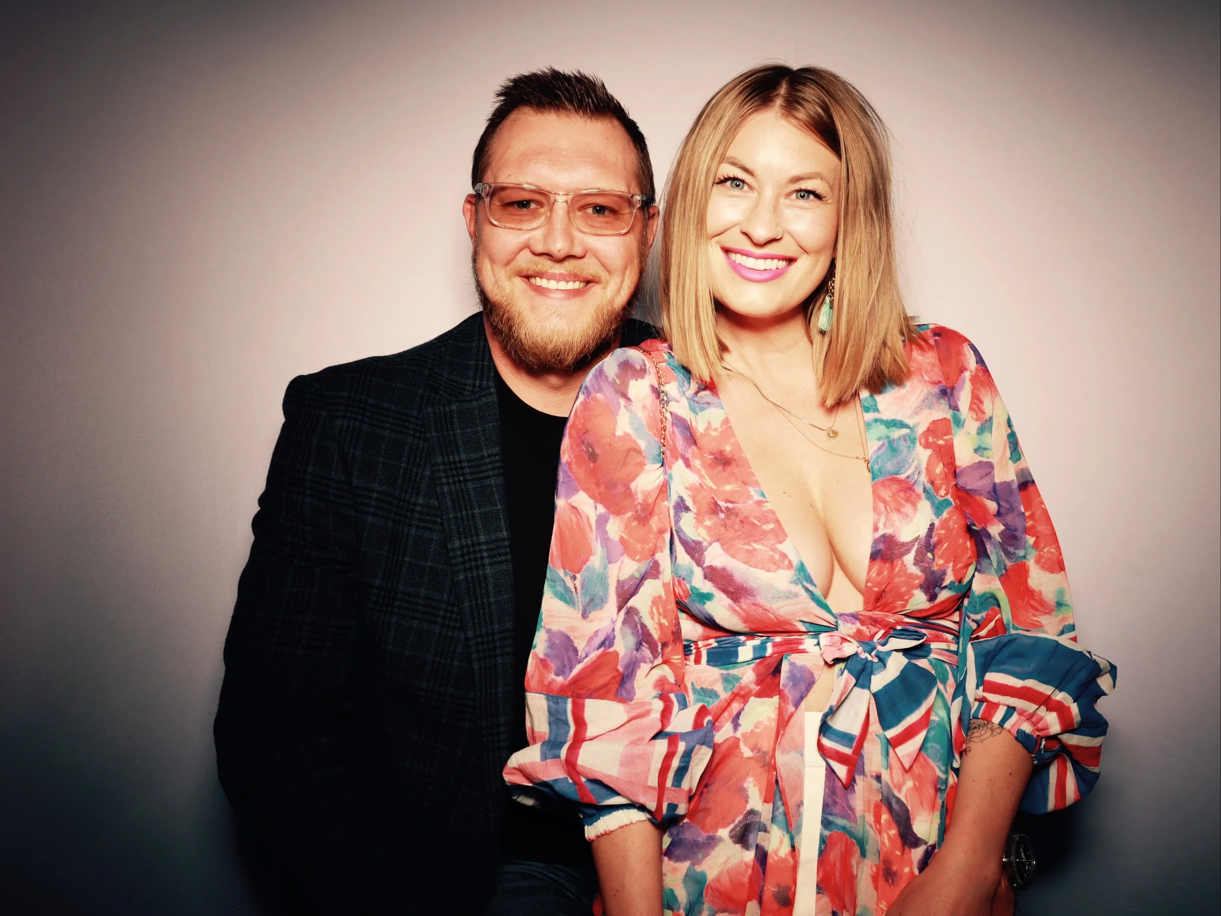 A smiling man with glasses and a beard, and a smiling woman with shoulder-length blonde hair, wearing a colorful floral dress with a deep neckline, standing against a plain background.