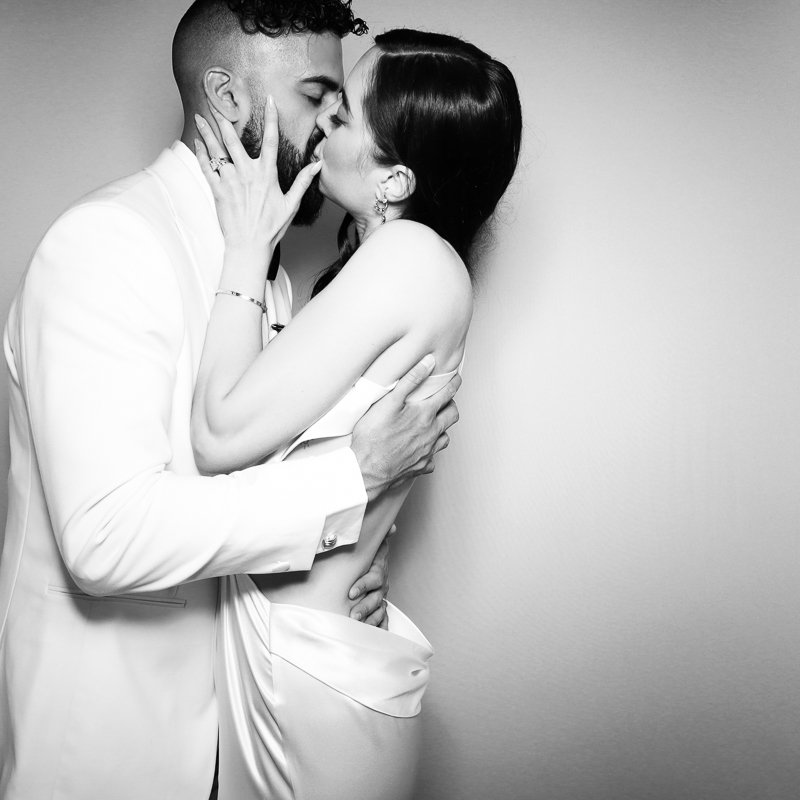 A black-and-white photo of a man and woman sharing a kiss. The man is wearing a white suit, and the woman has dark hair and is dressed in a satin gown. They are embracing each other closely against a plain wall.