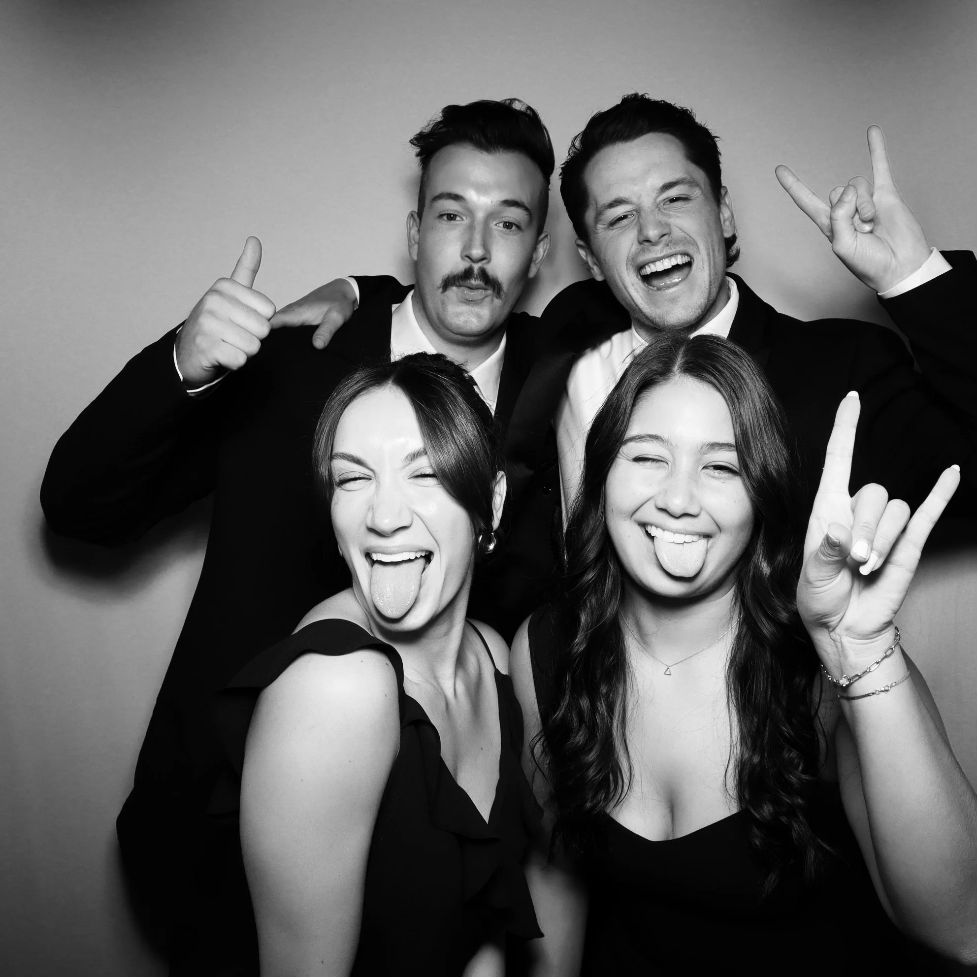 Four young adults at a social gathering, three men and two women, smiling and making playful gestures toward the camera, in black and white.