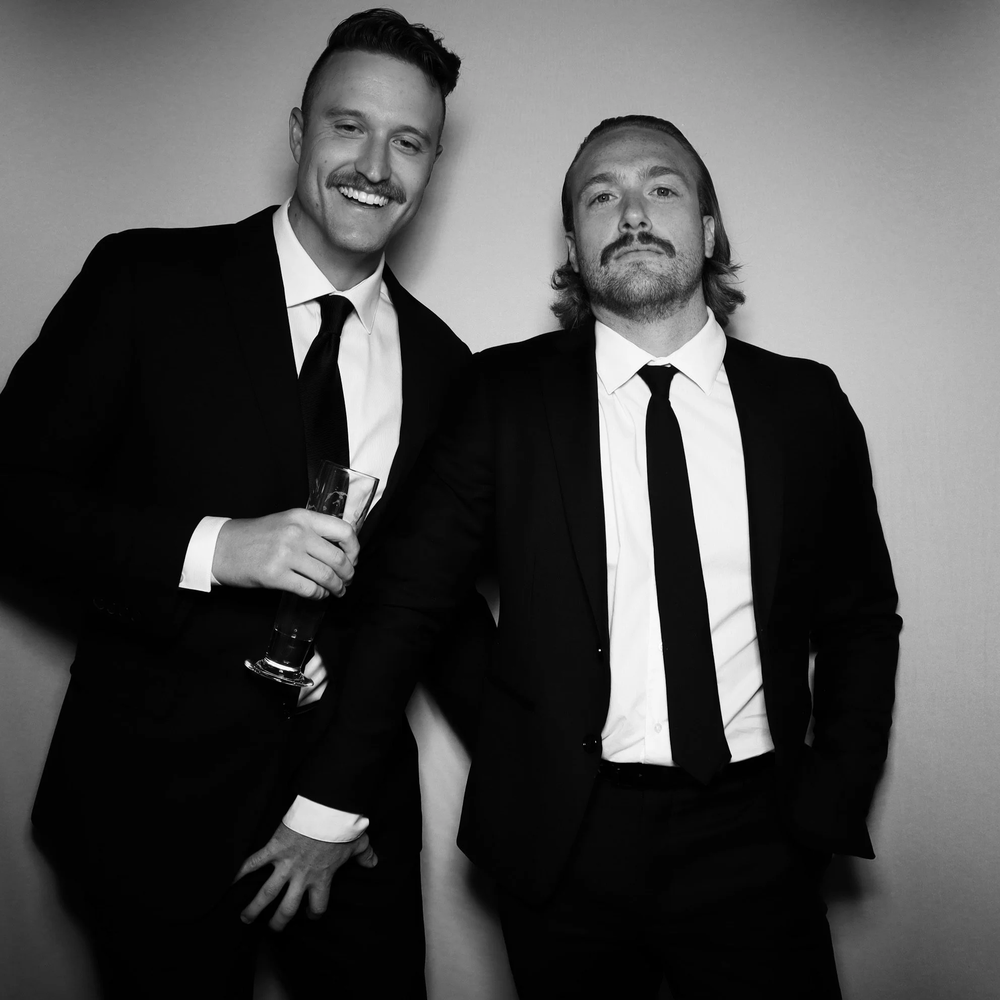 Two men in black suits, white shirts, and black ties standing together. The man on the left is holding a drink and smiling, while the man on the right has a serious expression with hands in pockets. Black and white photo.