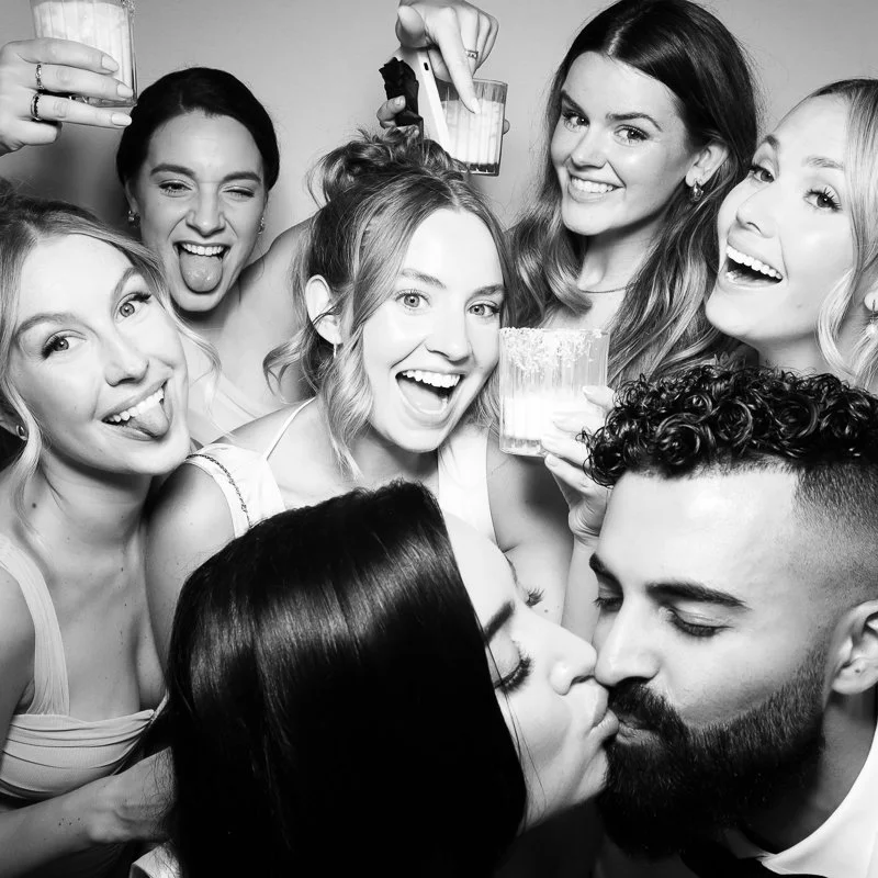 Group of excited young women at party, some making silly faces, one woman and a man kissing, enjoying drinks, in black and white.