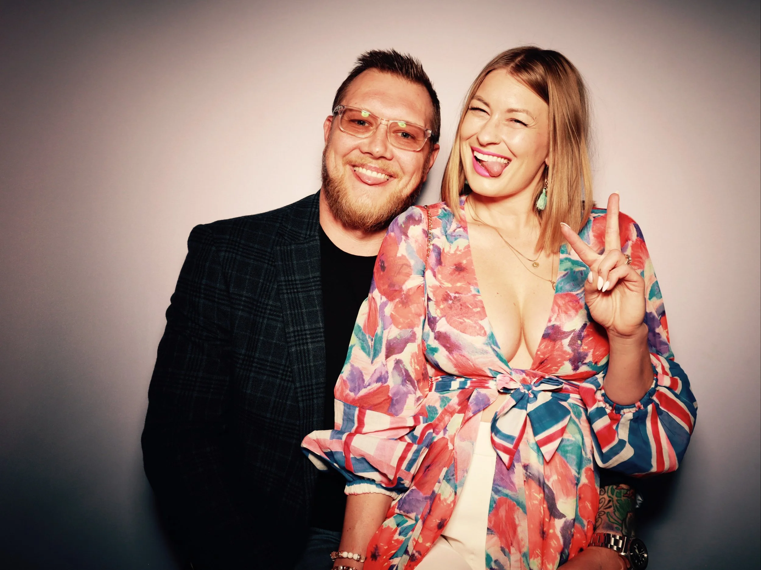 A man with glasses and a beard smiling and standing close to a woman making a peace sign and sticking out her tongue, both posing together against a plain background.