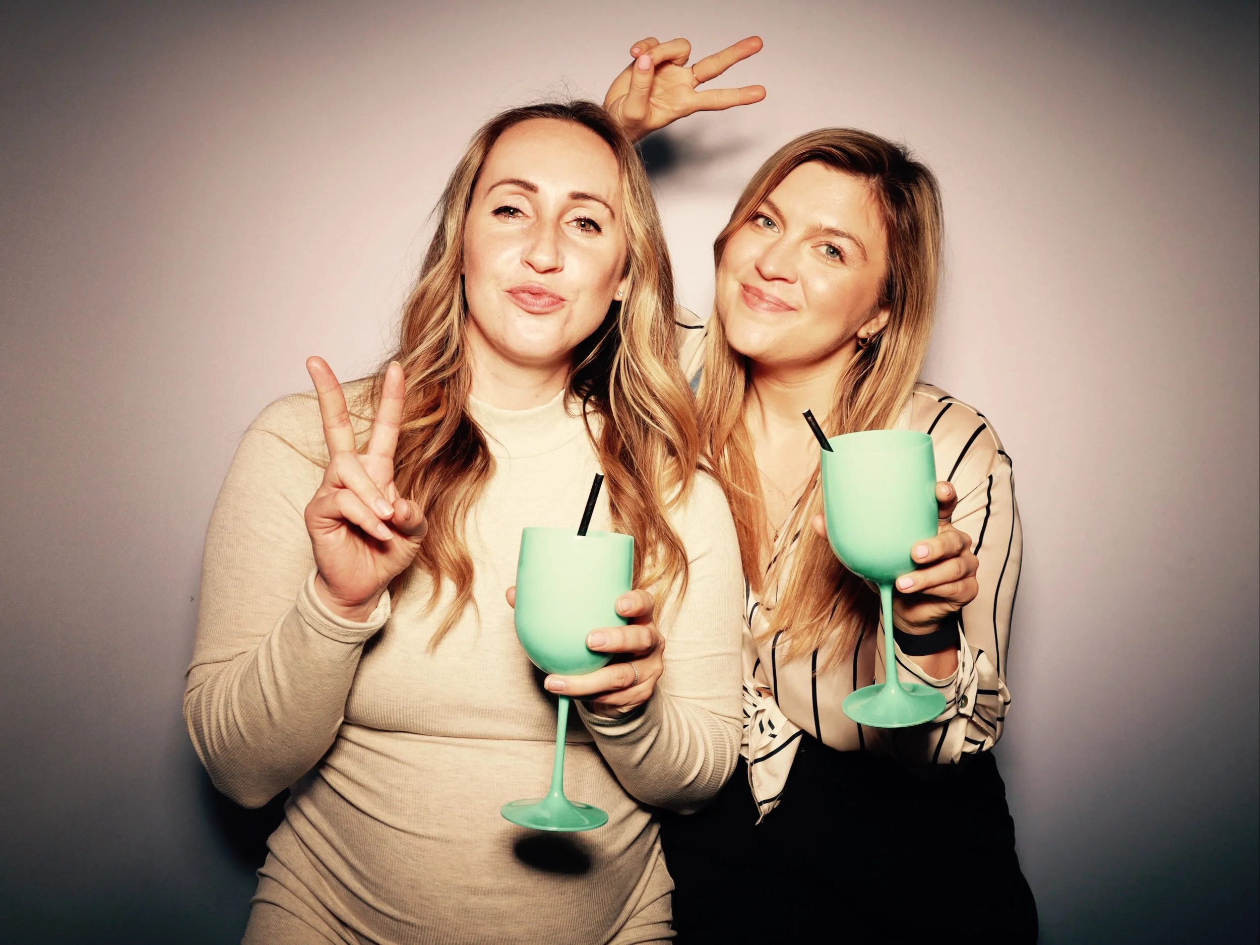 Two women smiling and posing for a photo, each holding a glowing mint green drink with a straw, one making a peace sign and the other raising a hand behind her head in a playful gesture.
