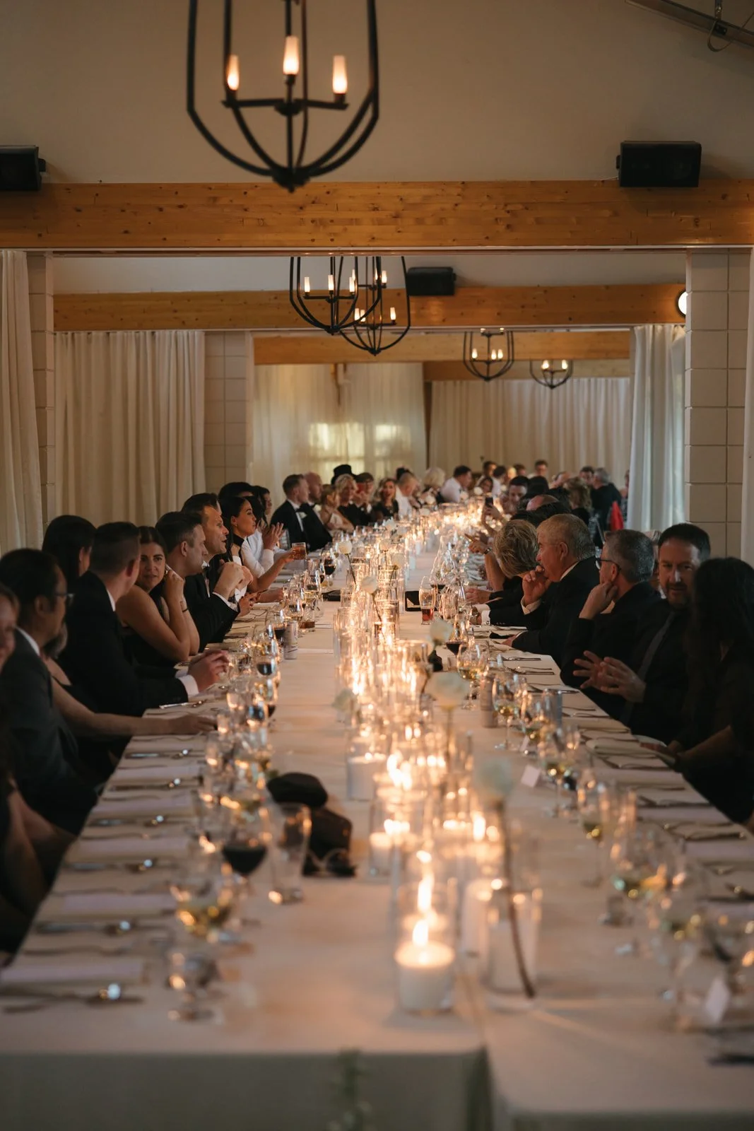 A long banquet table with people dressed in formal attire, dining, and socializing during an elegant event with warm lighting, candles, and a rustic interior.