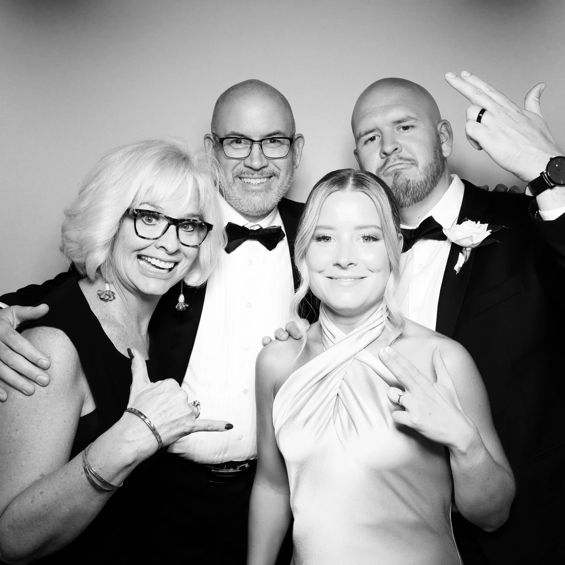 A group of five people dressed in formal attire posing together for a photo, some making hand gestures, with a plain background.