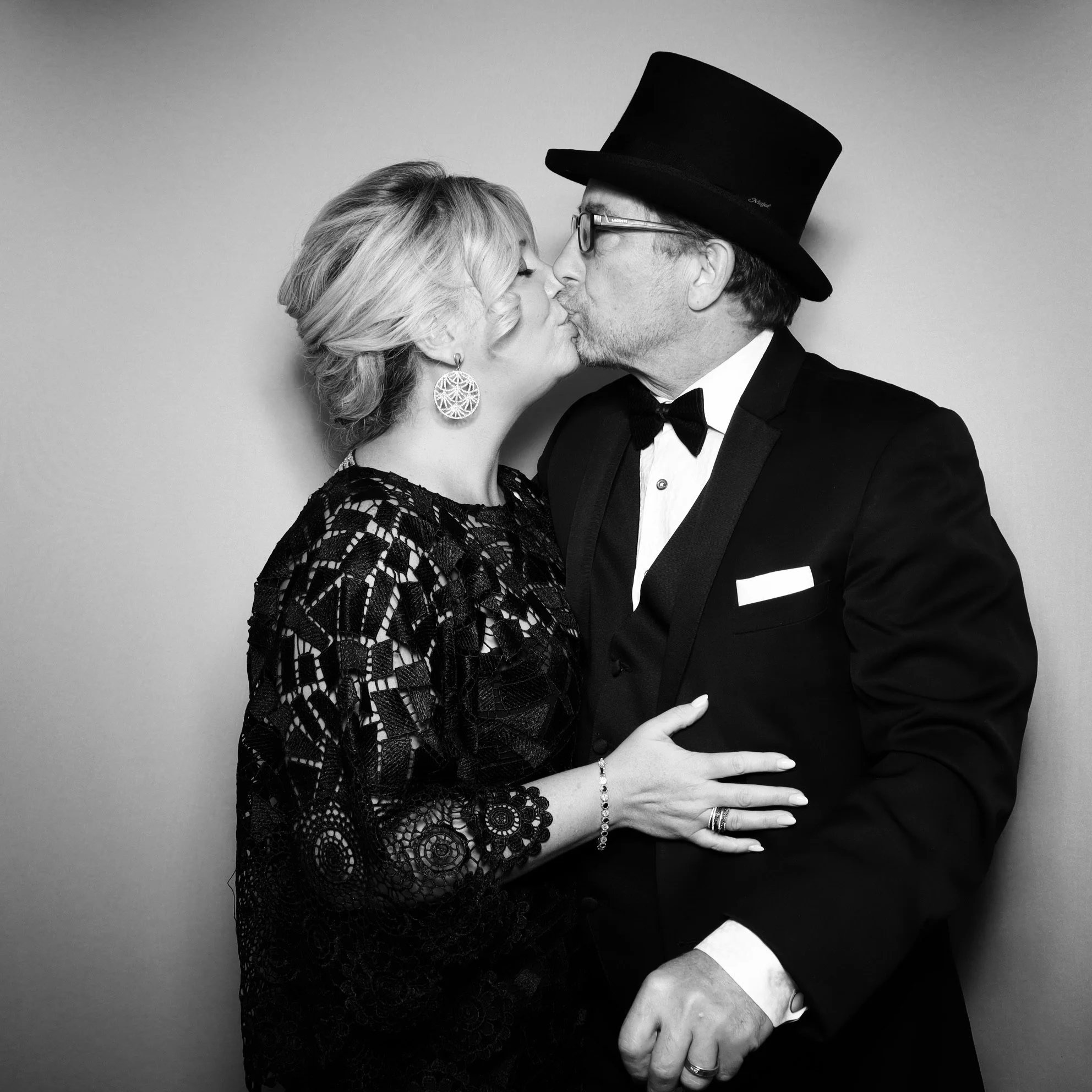 A man and woman kissing, both dressed in formal attire, with the man wearing a top hat and glasses, against a plain background.