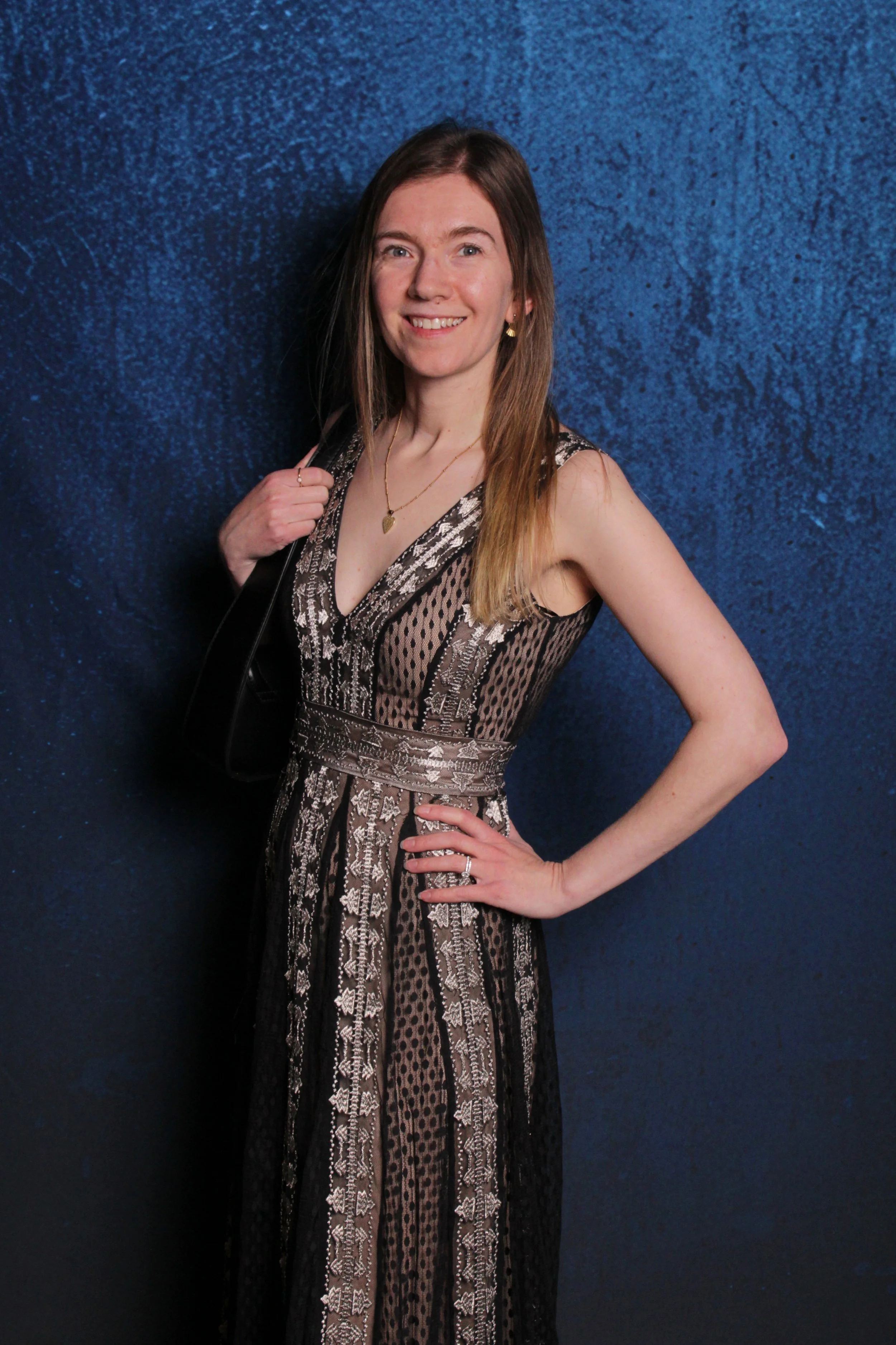 A woman with long hair wearing a black and silver patterned dress, smiling, standing against a blue textured background.