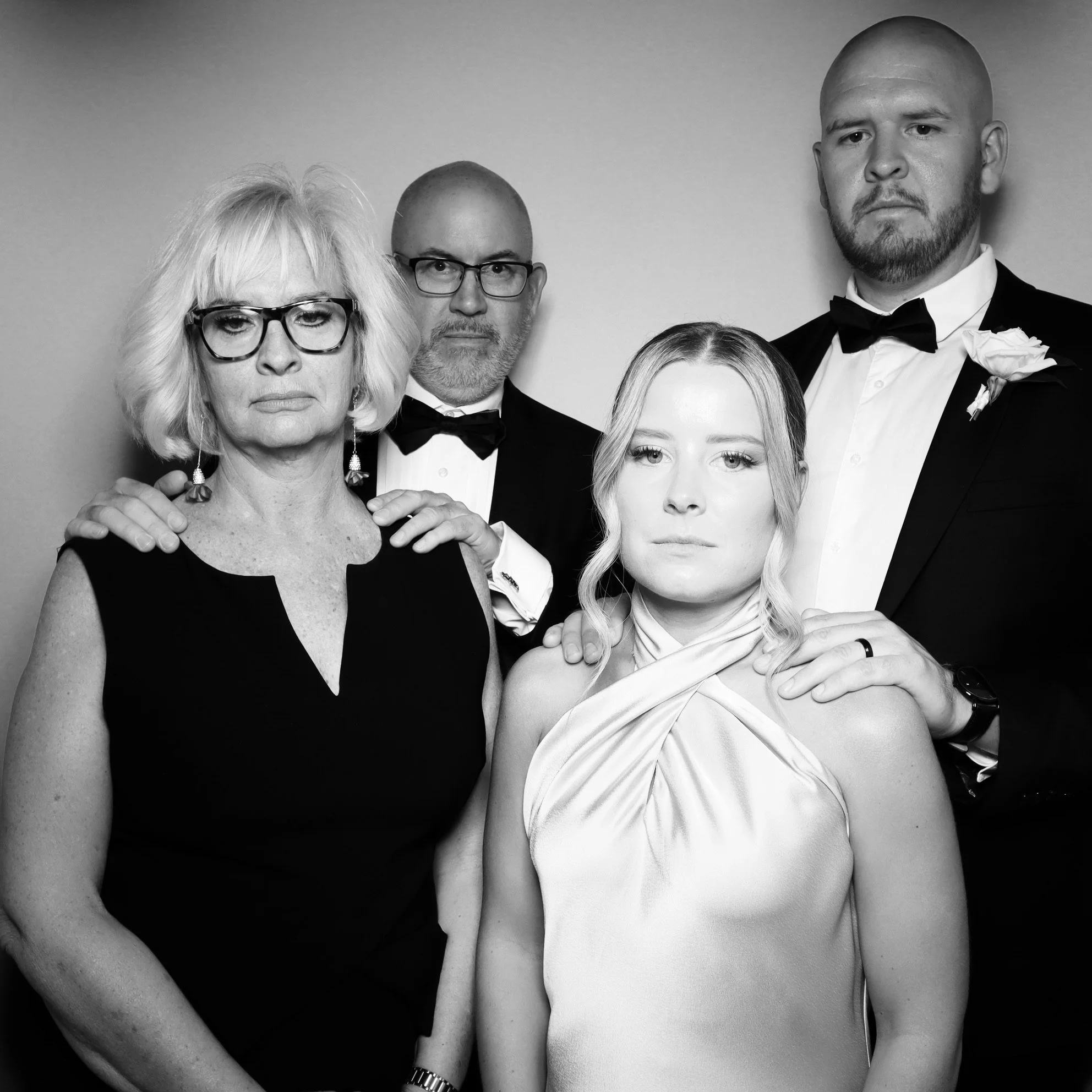 A black-and-white portrait of five people dressed in formal attire, with three men in tuxedos and two women, one older with glasses and a younger woman, standing close together.