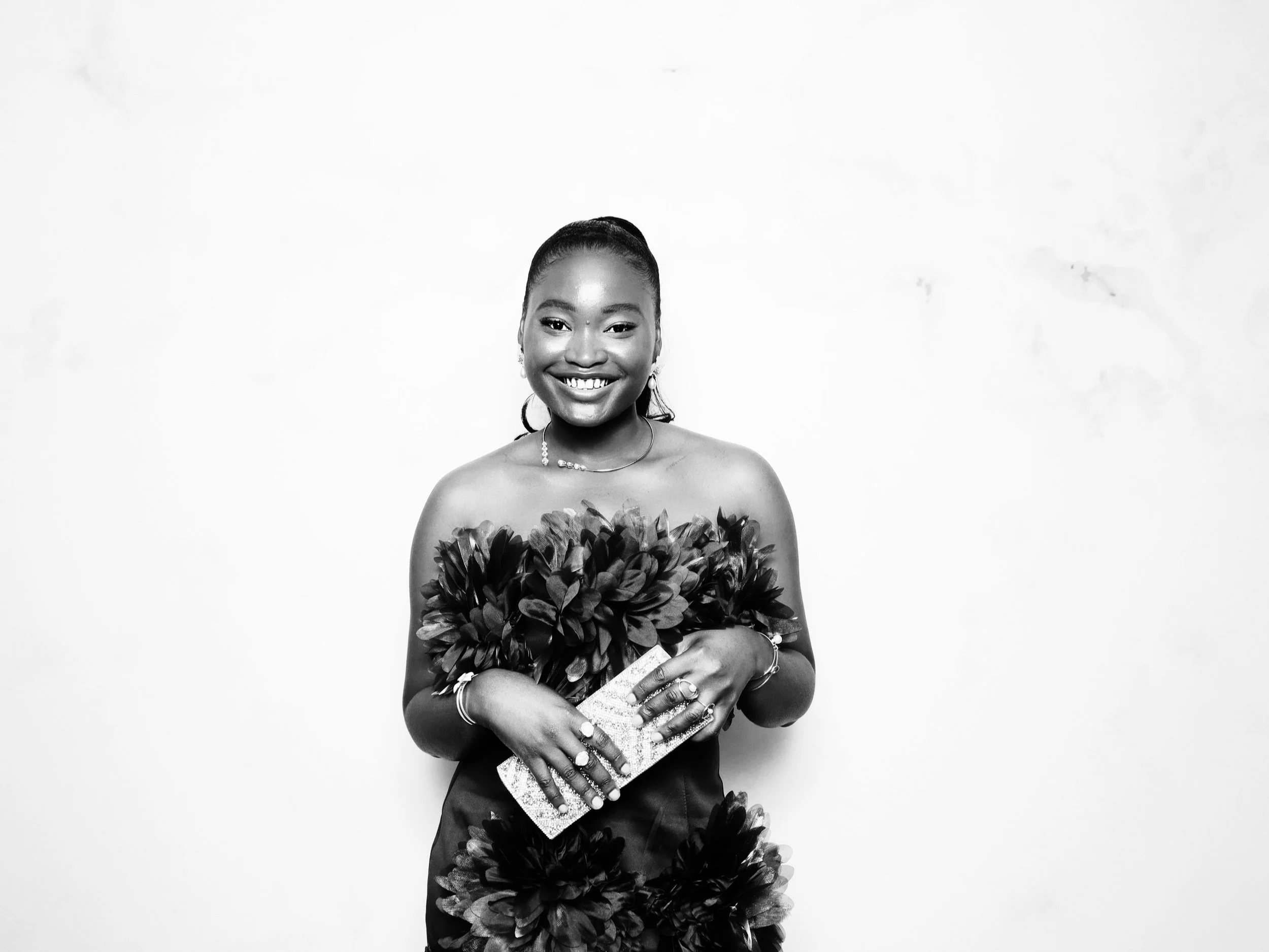 A young woman in a strapless dress with large floral embellishments is smiling and holding a clutch purse, standing against a plain white wall.