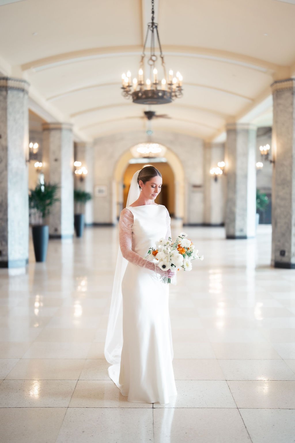 A bride in a white gown with lace sleeves holding a bouquet of white and orange flowers stands in a spacious, elegant hallway with chandeliers and tall columns.