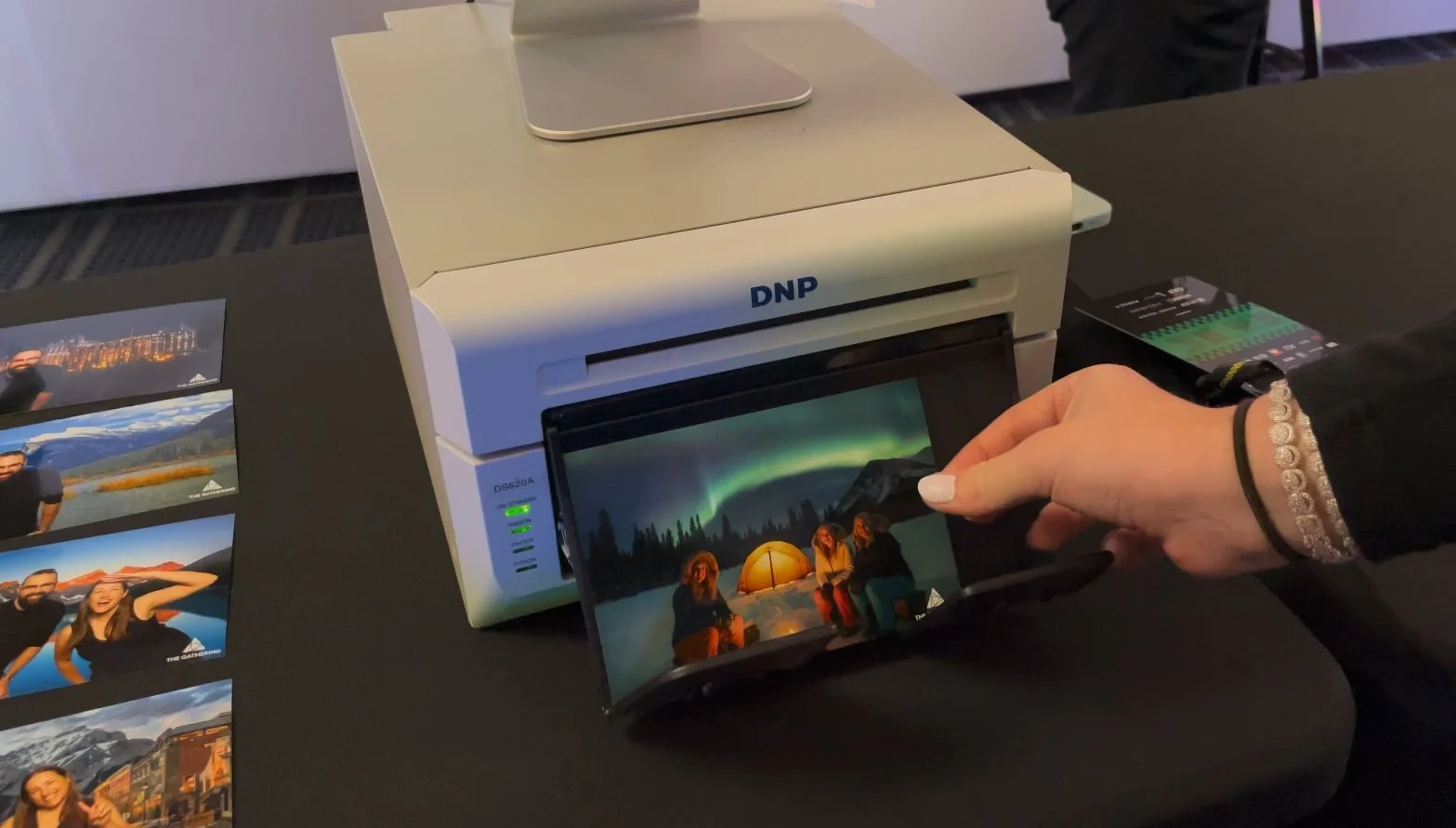 A person places a photo into a photo reduction machine. The photo depicts people sitting around a campfire near a tent at night with the northern lights overhead.