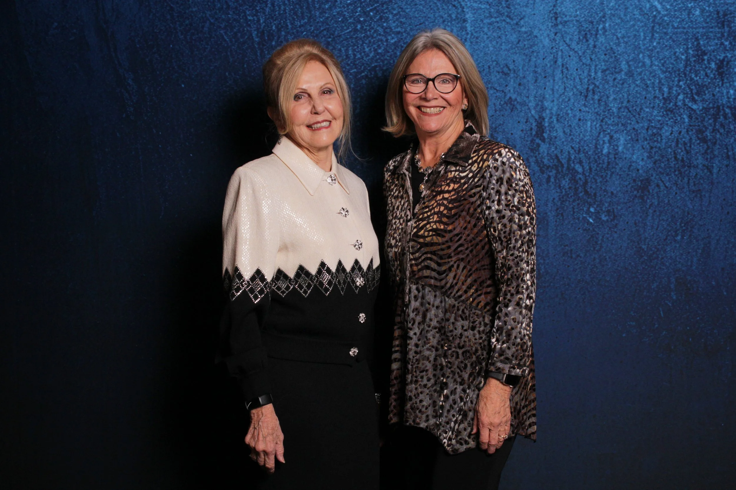 Two women smiling, standing with their arms close together against a dark blue textured background.