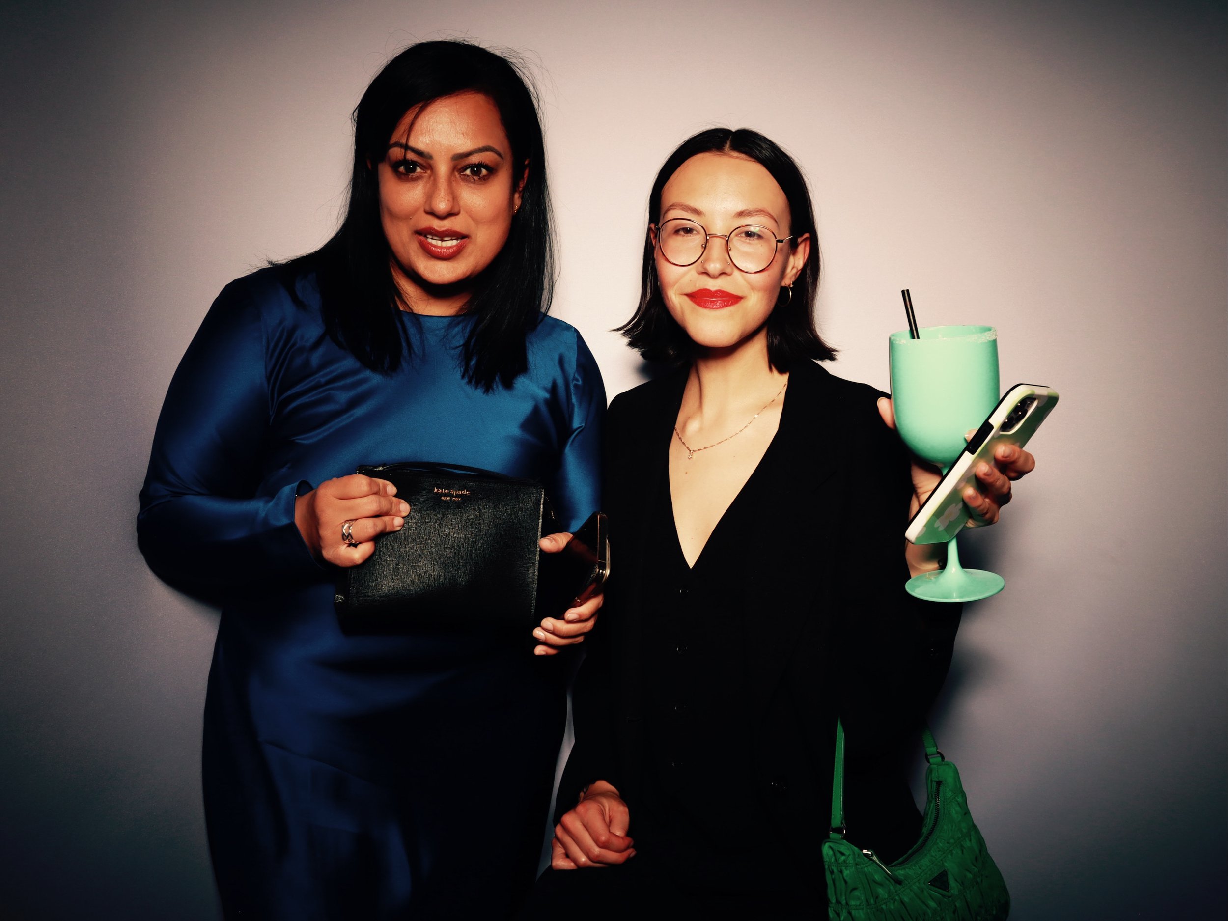 Two women standing together against a plain background, one holding a black purse and the other holding a large green cocktail with a phone in her hand.