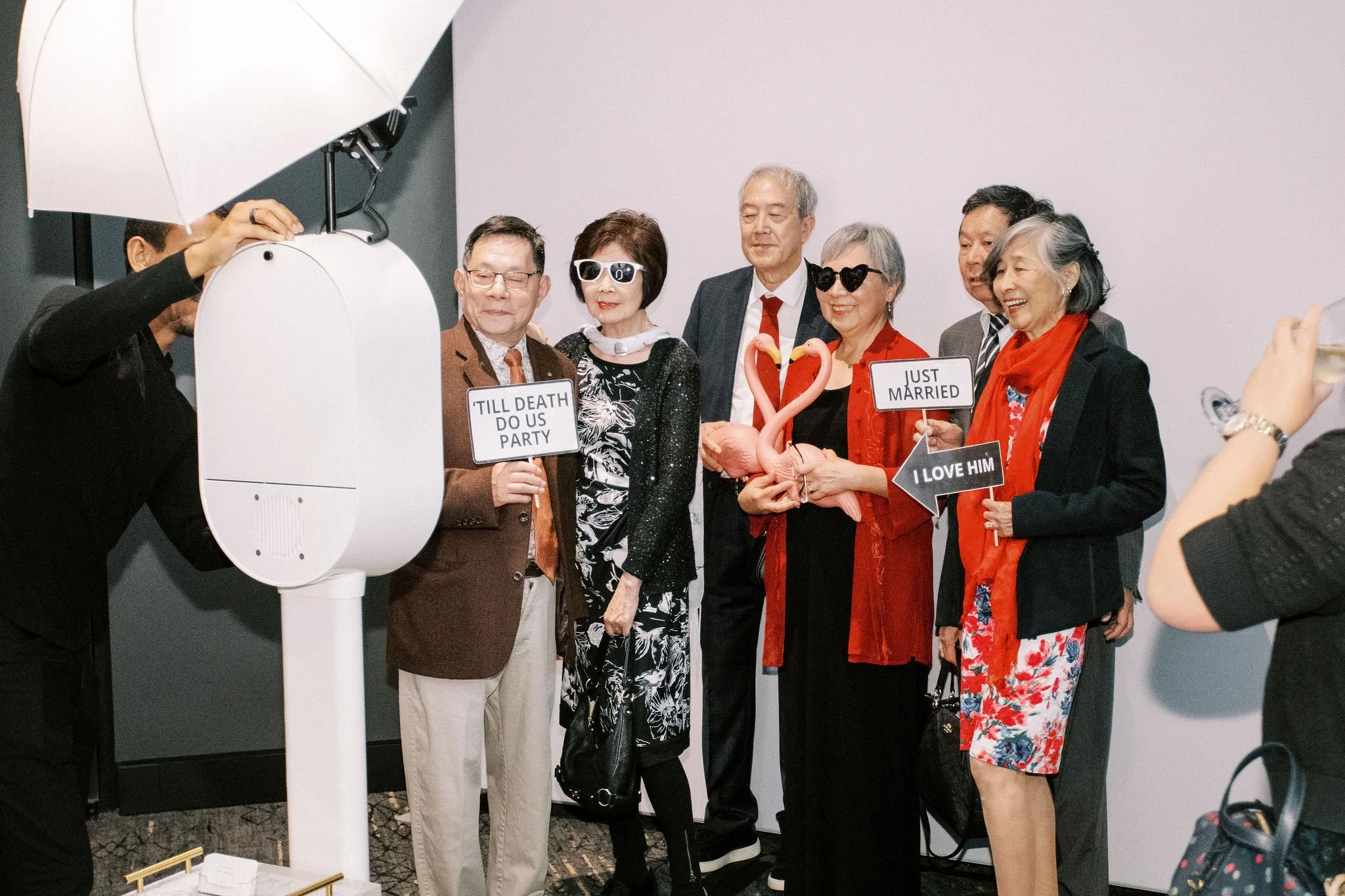 Group of elderly people at a wedding photo booth, holding signs that read 'Till Death Do Us Party,' 'Just Married,' and 'I Love Him,' with some wearing sunglasses and props including pink flamingos, in a festive, indoor setting.