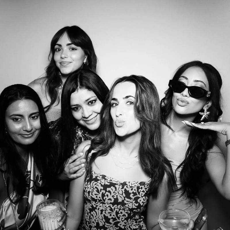 Five women taking a group photo, some smiling and others making playful expressions, in a black and white setting.