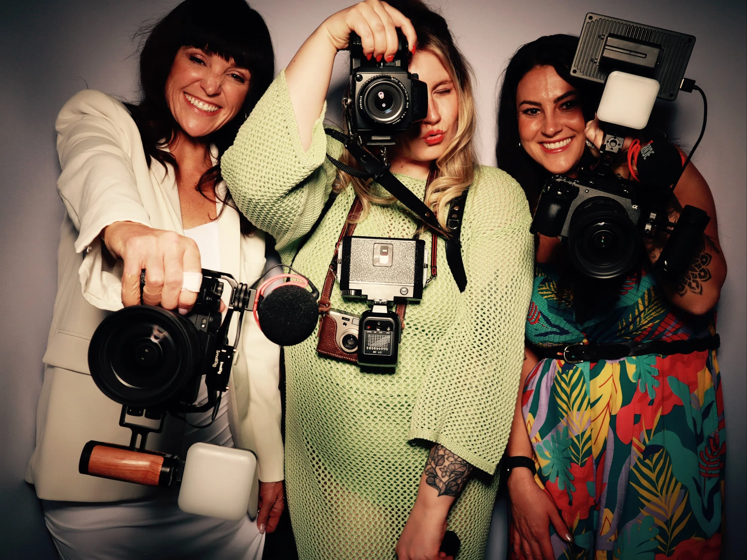 Three women holding professional cameras and photography equipment, smiling and posing together in front of a gray background.