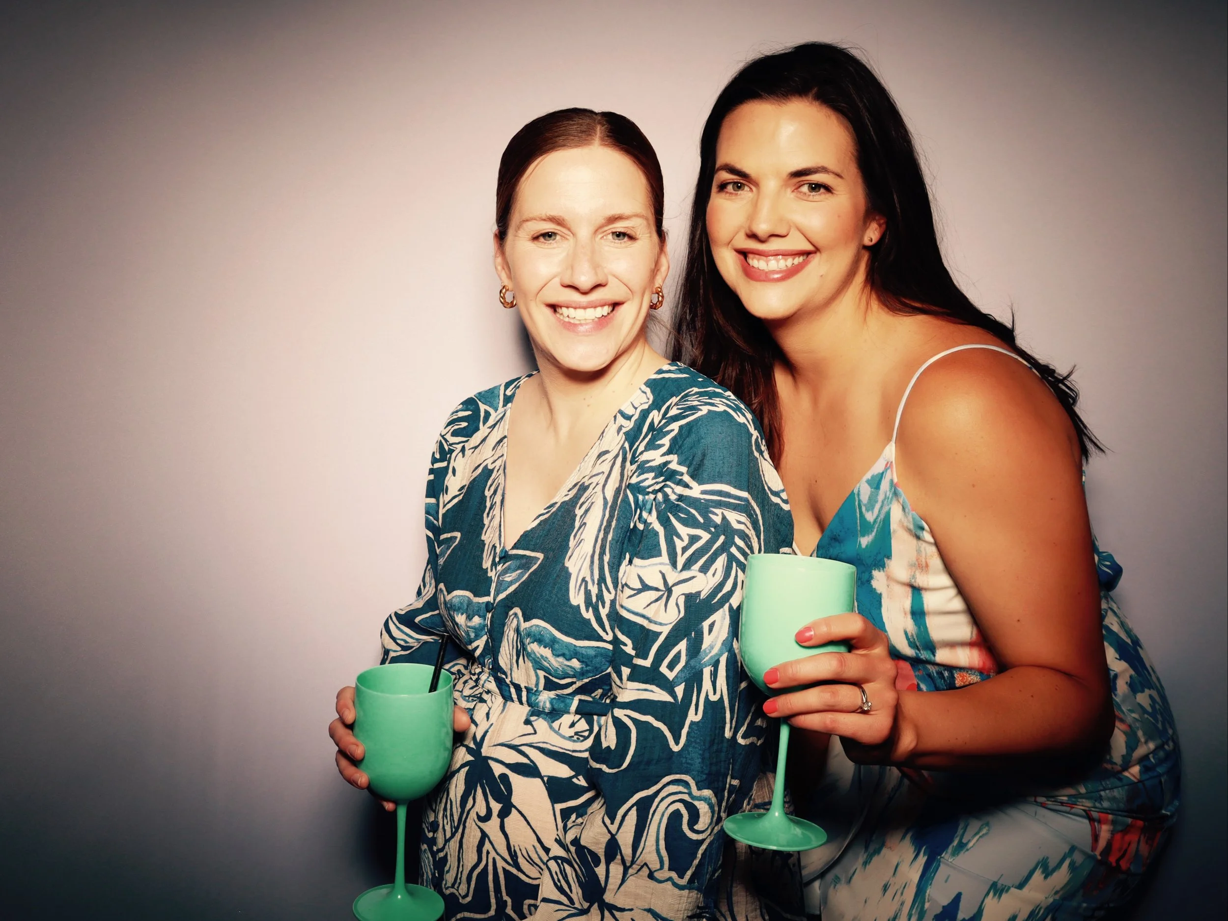 Two women smiling and holding green wine glasses with straws, standing close together against a plain background