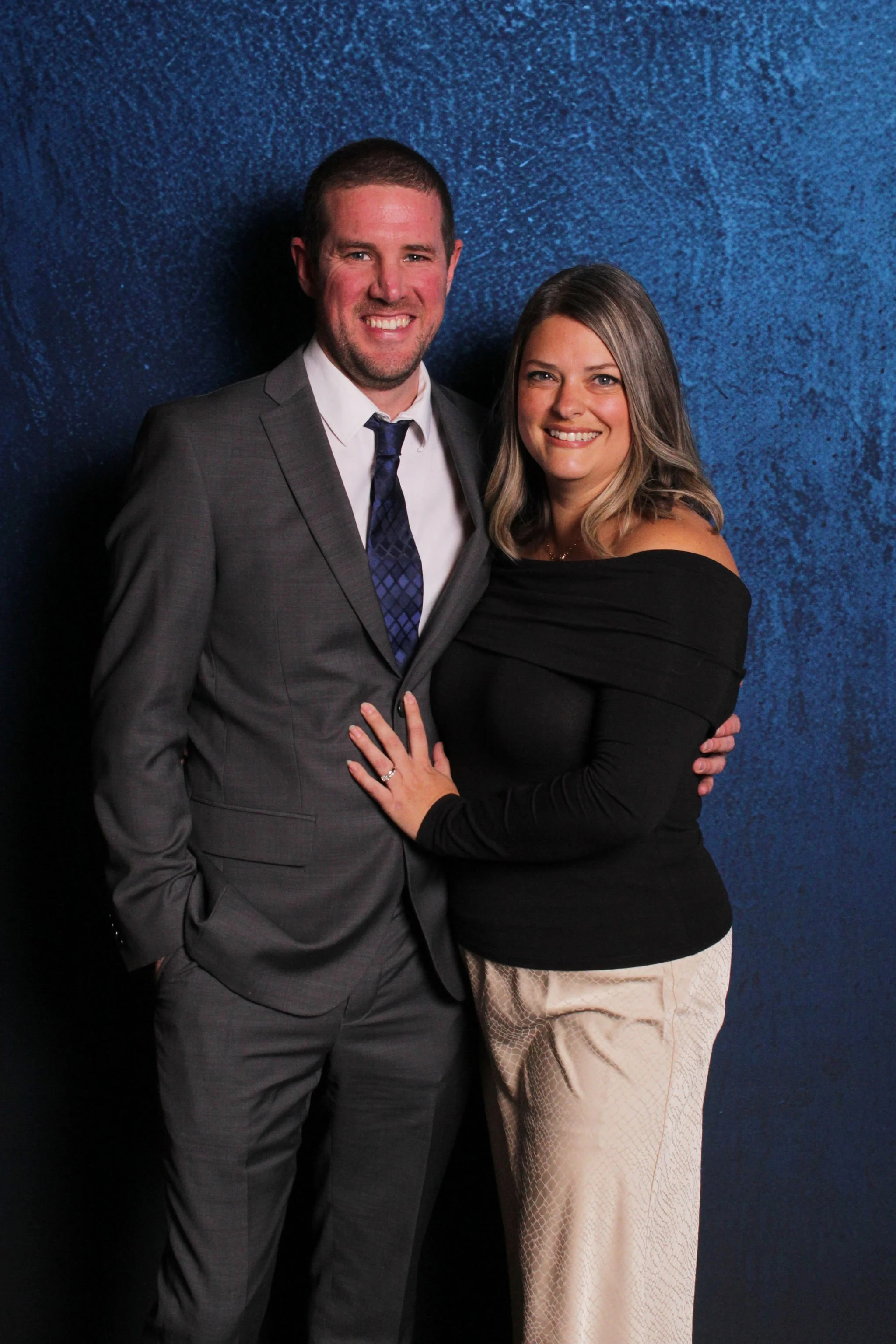 A man in a gray suit and woman in a black off-shoulder top pose together against a blue textured background, smiling