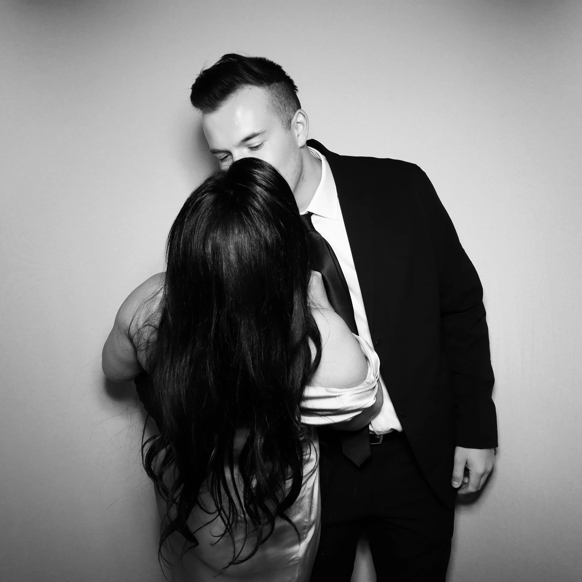 A man and woman with dark hair are close together, with their foreheads touching, against a plain wall. The man is wearing a suit and tie, and the woman is in a dress.