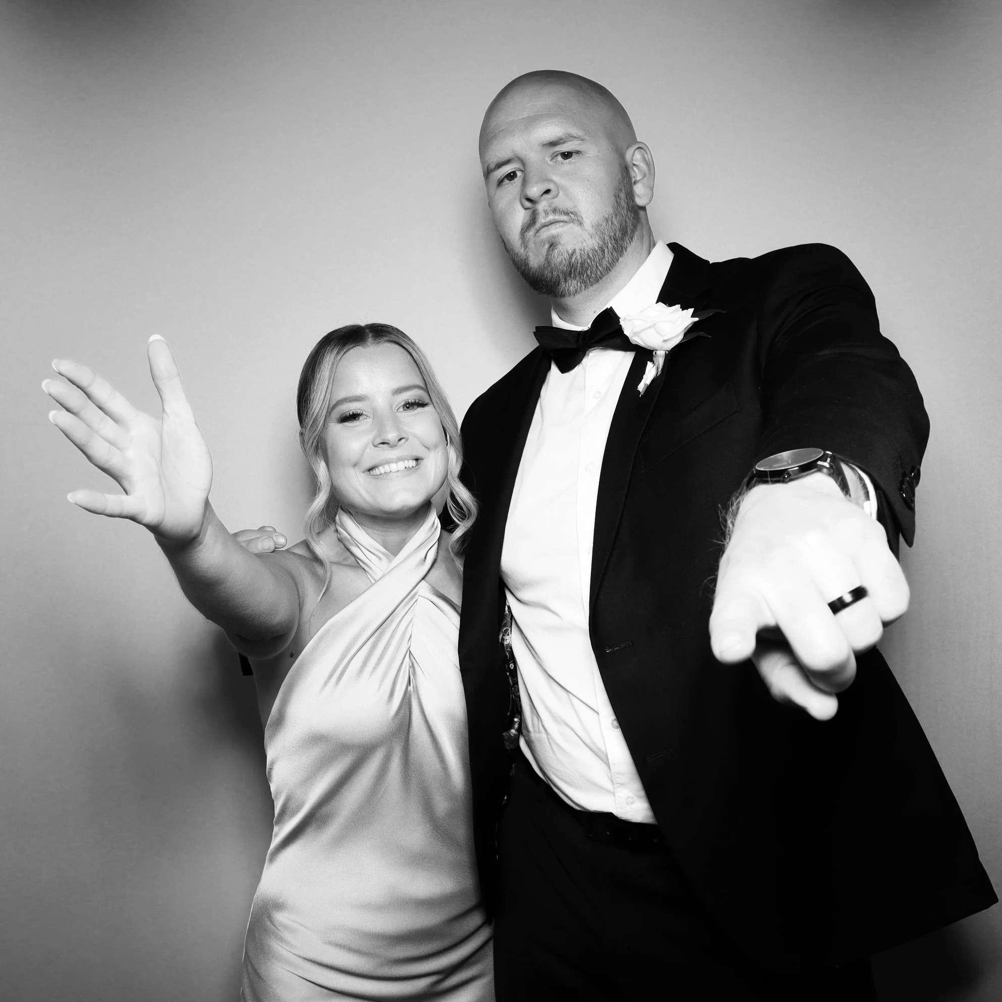 A woman and a man in formal attire, with the man wearing a tuxedo and the woman in a satin dress, posing playfully for a photo with a plain background.
