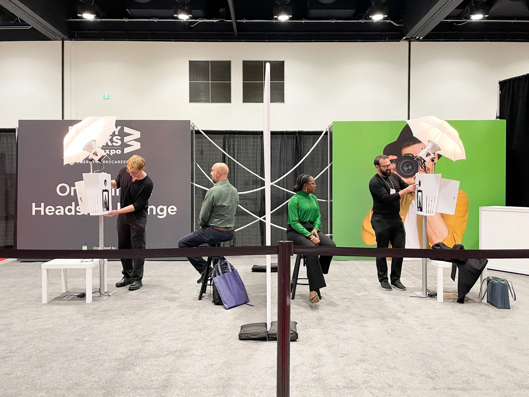 At a career fair or expo, three individuals are sitting or standing next to a booth that features a large backdrop with a person holding a camera to their eye. There are two umbrellas with lights on each side of the backdrop. One person appears to be working with a display, another is seated with a green shirt, and a third is standing with a beard and glasses. The booth has signage related to career exchange or headshots.