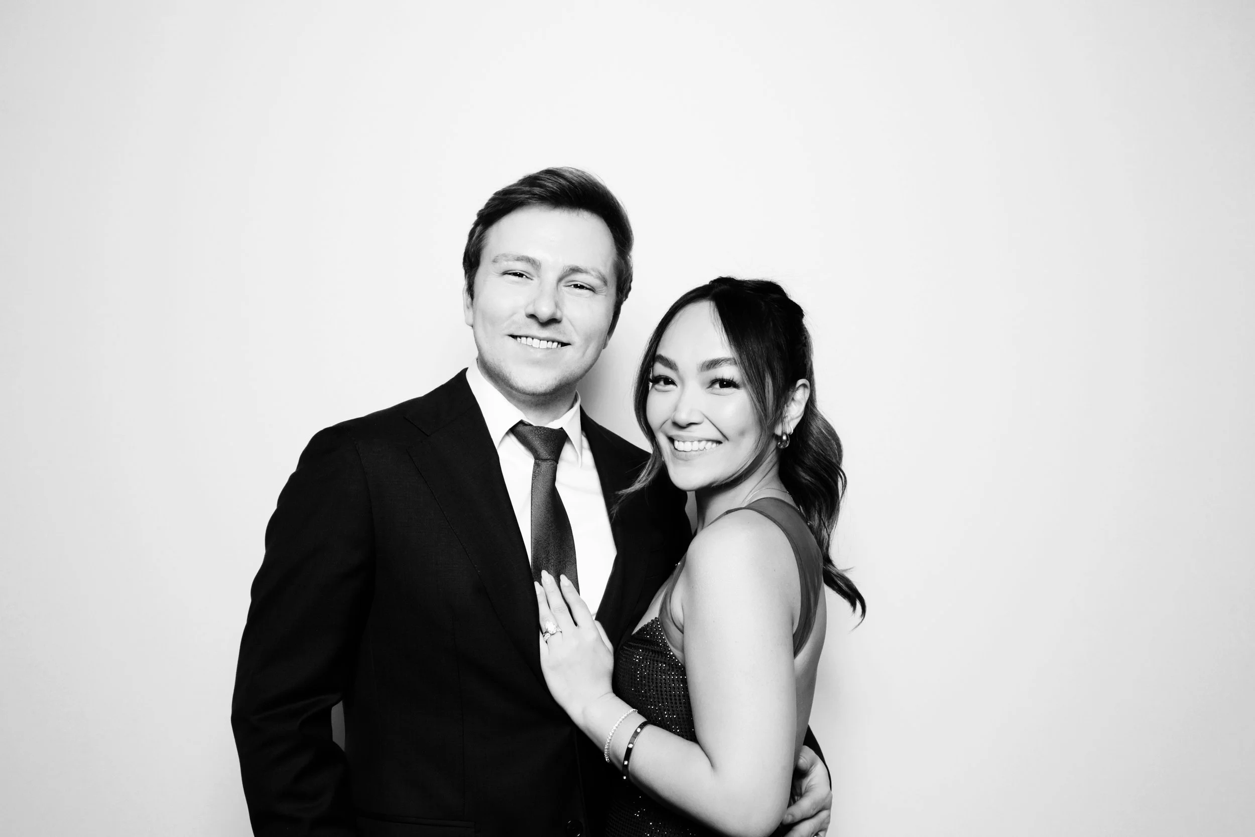 A black-and-white photo of a smiling couple in formal attire, standing close to each other against a plain background.
