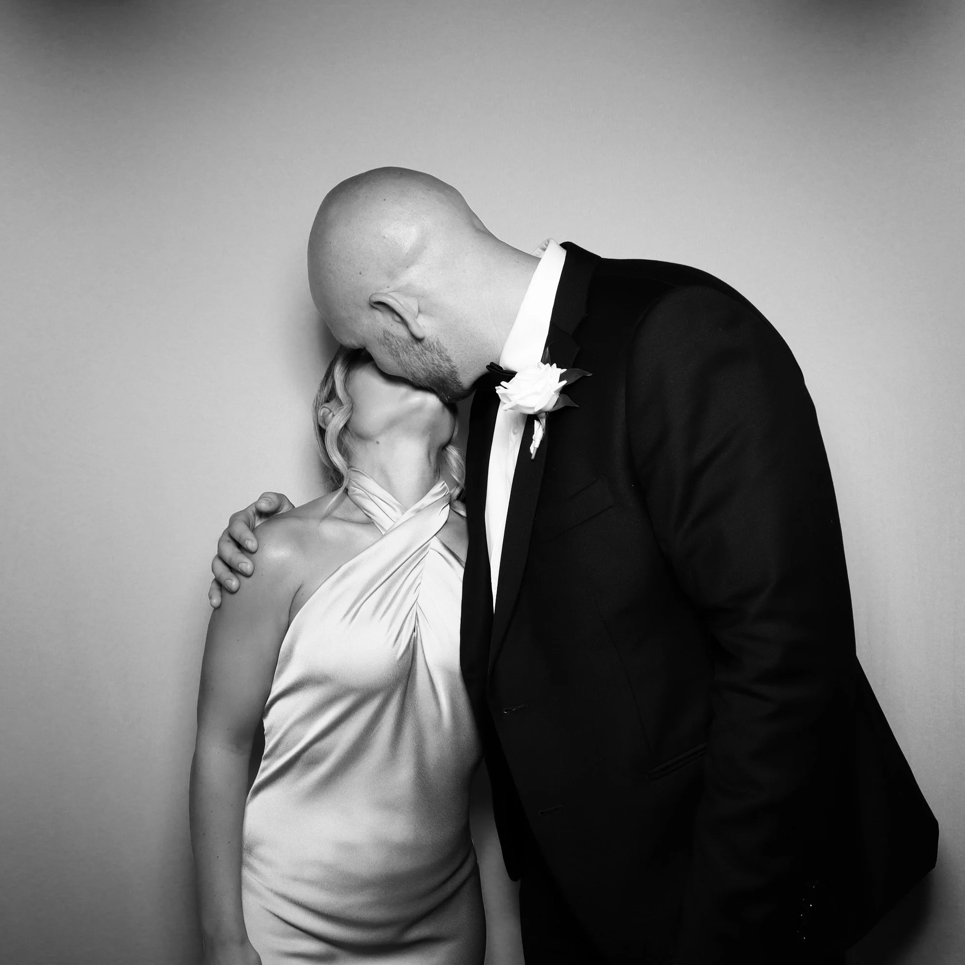A black and white photo of a man and woman kissing, with the man dressed in a tuxedo and the woman wearing a satin dress.
