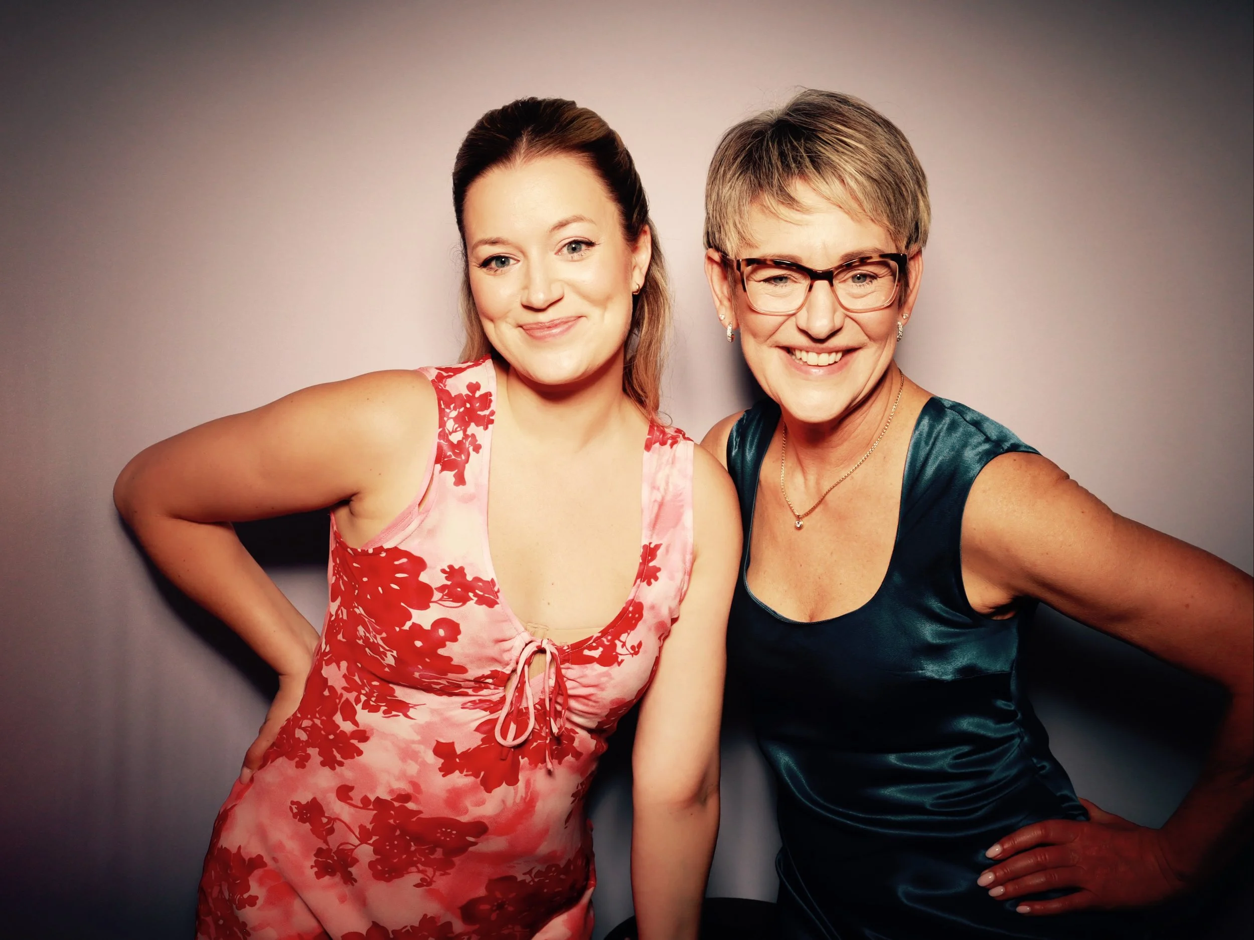 Two women posing together, smiling, against a plain background. The woman on the left is wearing a pink and red floral sleeveless dress, and the woman on the right is wearing a dark teal sleeveless dress with glasses.