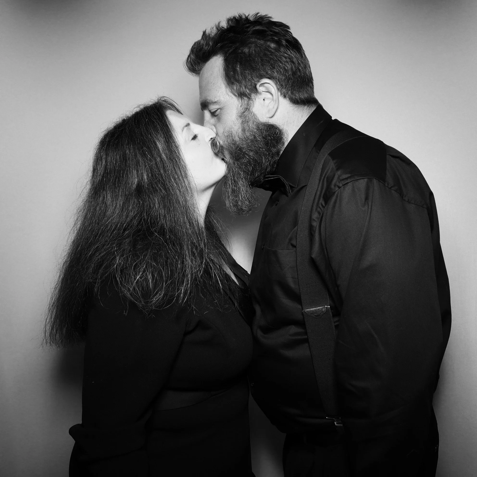Black and white photo of a woman and a man with a beard sharing a close, intimate moment, about to kiss, against a plain background.