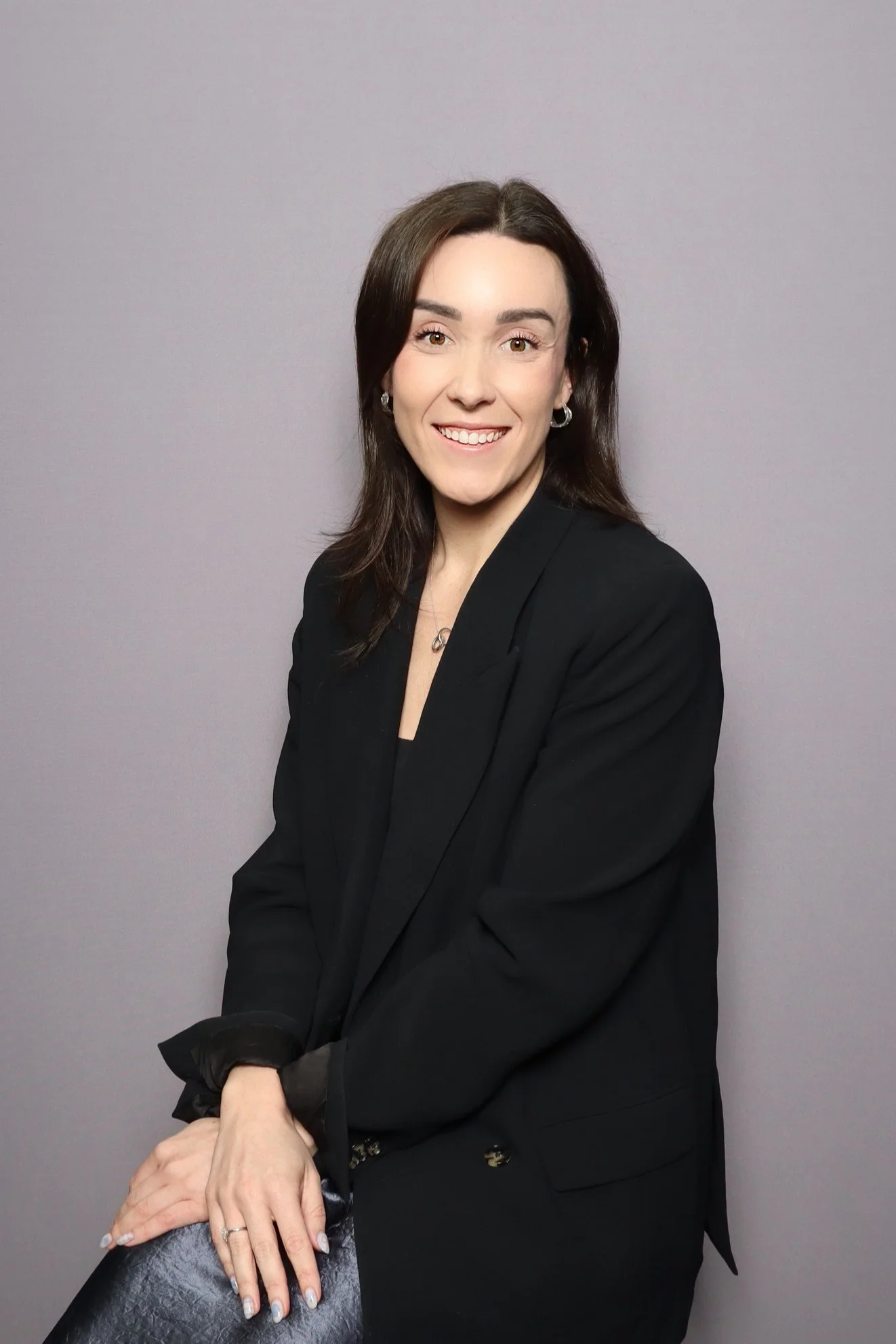 A woman with brown hair wearing a black blazer, smiling, sitting on a black chair against a gray background.