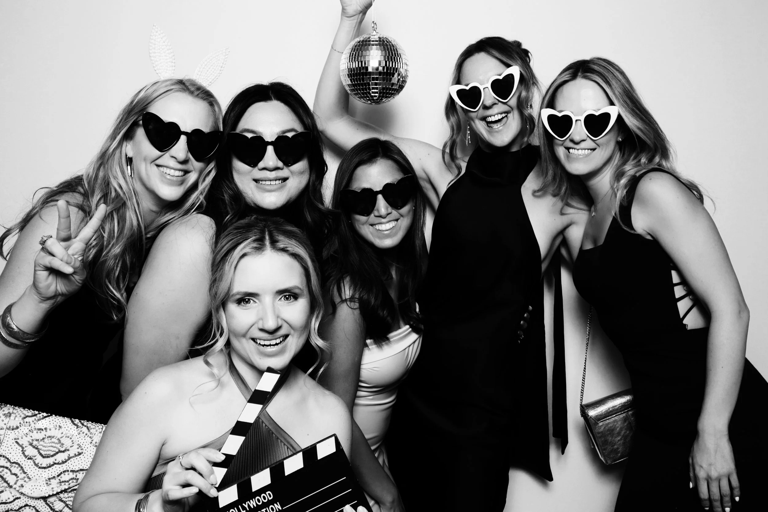 Group of seven women at a party, smiling and wearing heart-shaped sunglasses, with one woman holding a clapperboard. They are posing in front of a plain background, some making peace signs. One woman holds a disco ball overhead.