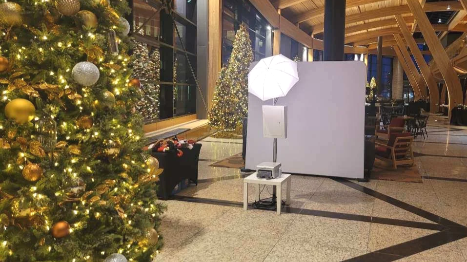 Indoor Christmas scene with decorated Christmas trees, a photo booth setup with a white backdrop, a large umbrella light, and tables and chairs in the background.