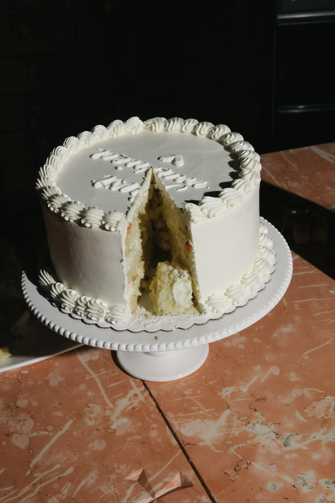 White frosted birthday cake with a slice removed, placed on a white cake stand on a marble countertop.