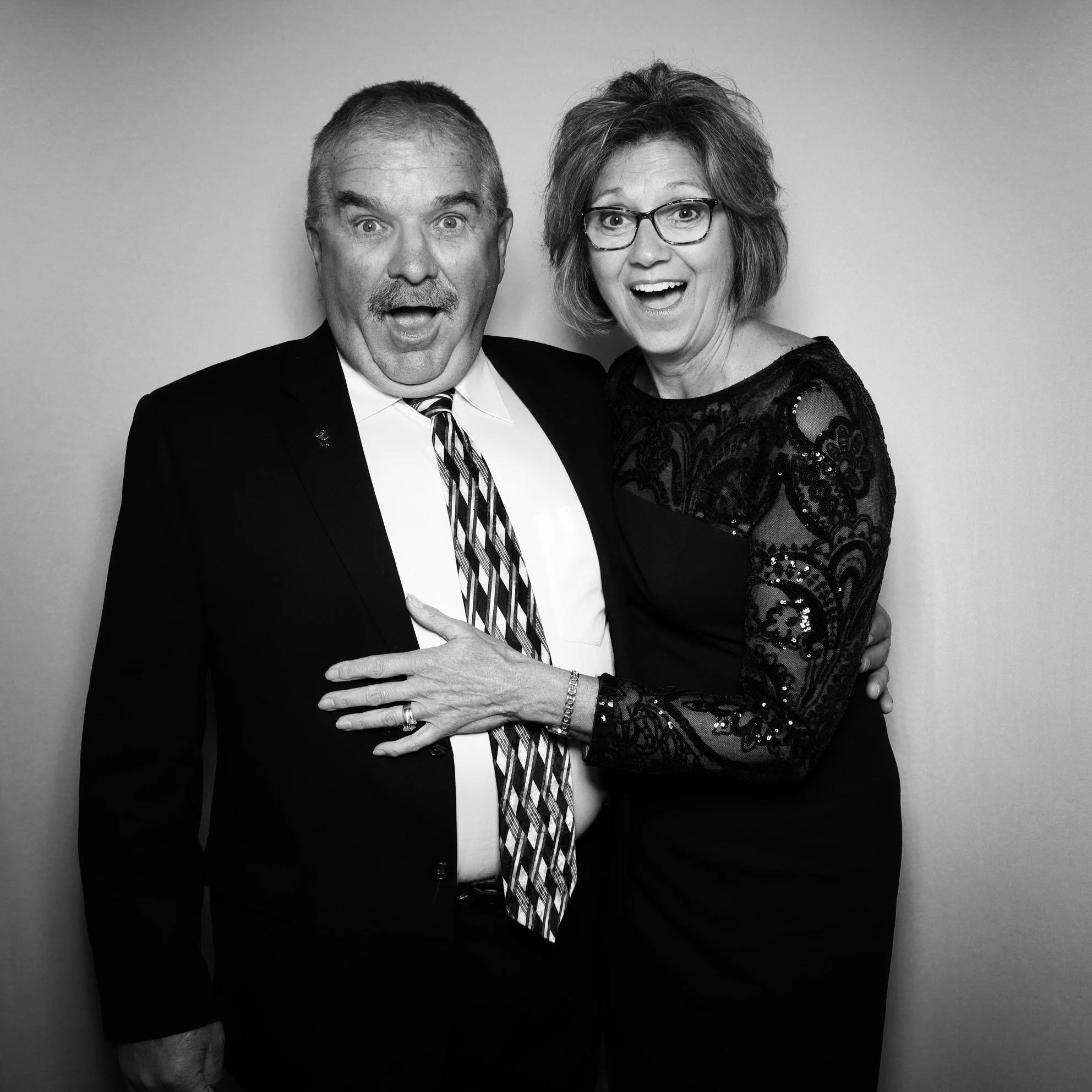 A black and white photo of a man and woman posing together, both with surprised expressions. The man is wearing a suit and tie, and the woman is wearing glasses and a dress with lace details.