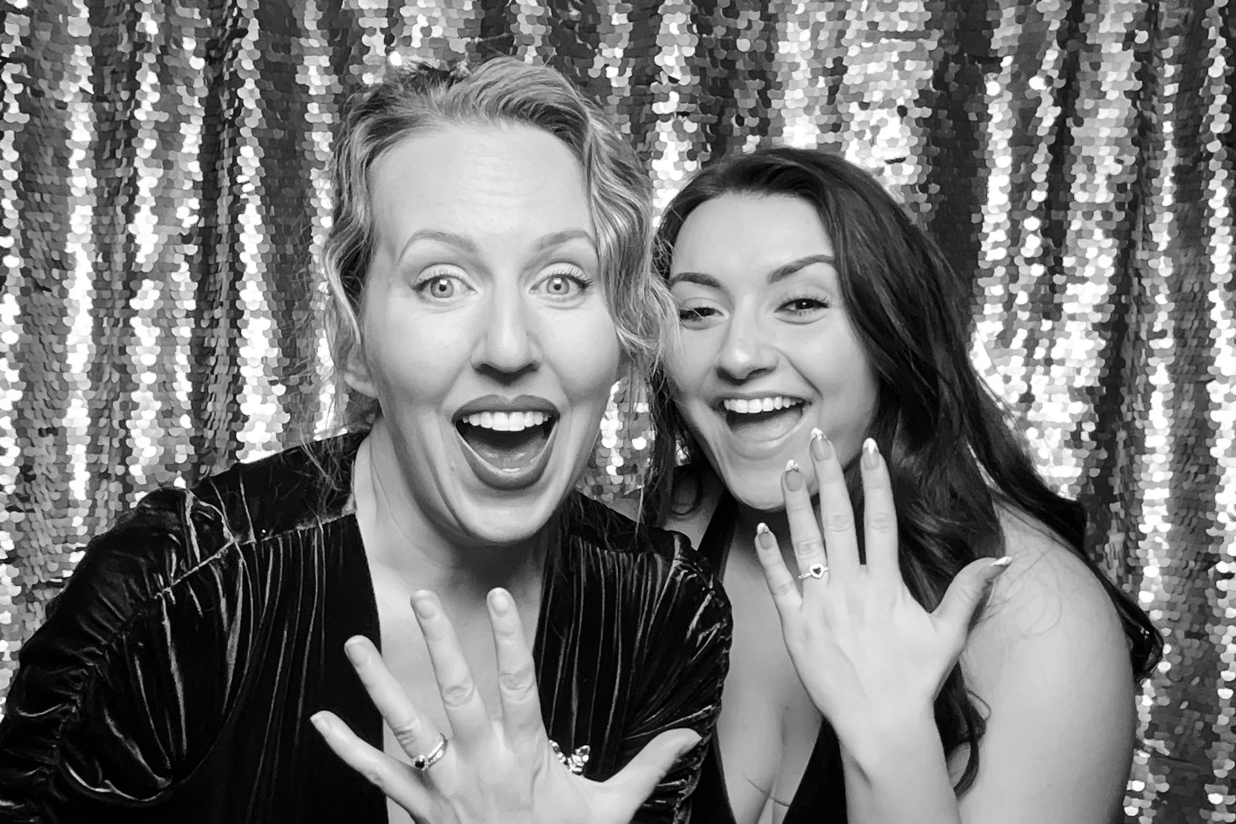 Two women smiling and showing off rings on their fingers in front of a sequin backdrop.