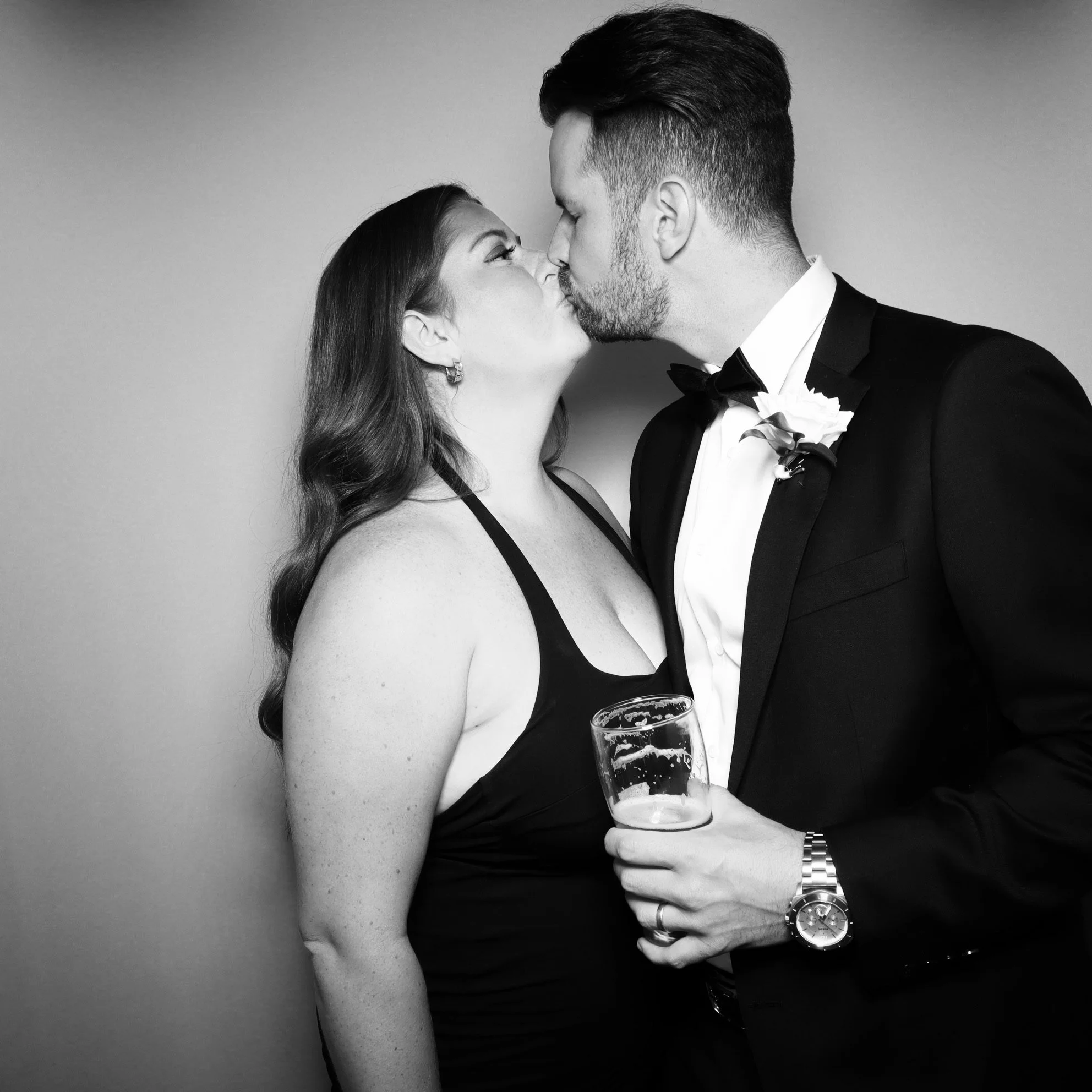A black and white photo of a couple in formal attire kissing, the woman wearing a black dress and the man in a tuxedo holding a glass of beer.