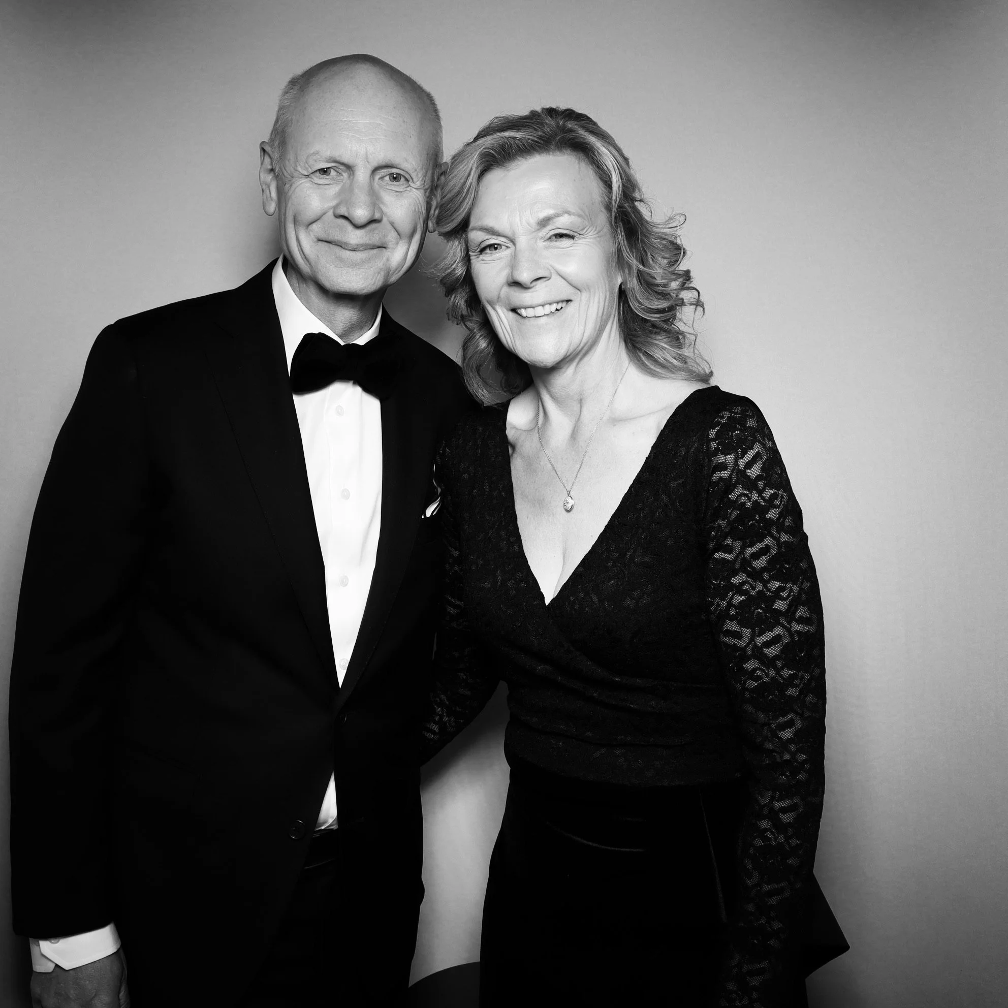 A black-and-white photo of an older man in a tuxedo and a woman in a black lace dress, smiling and posing together against a plain background.