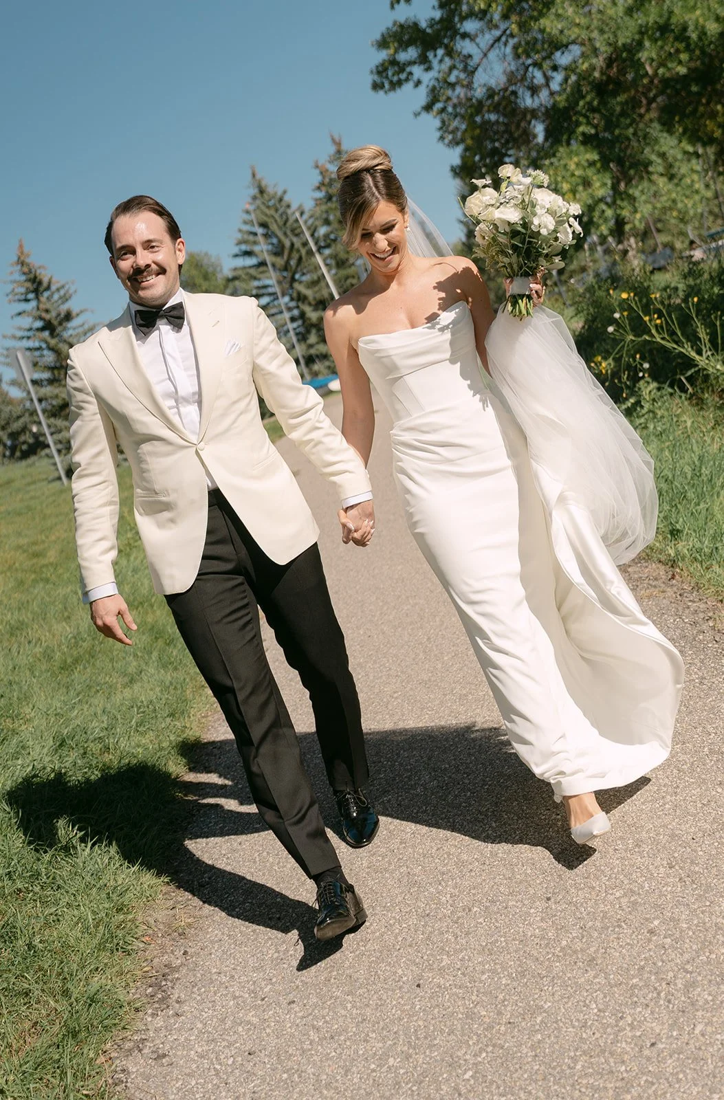 A bride and groom celebrating their wedding outdoors, holding hands and smiling, with green trees and a clear blue sky in the background.