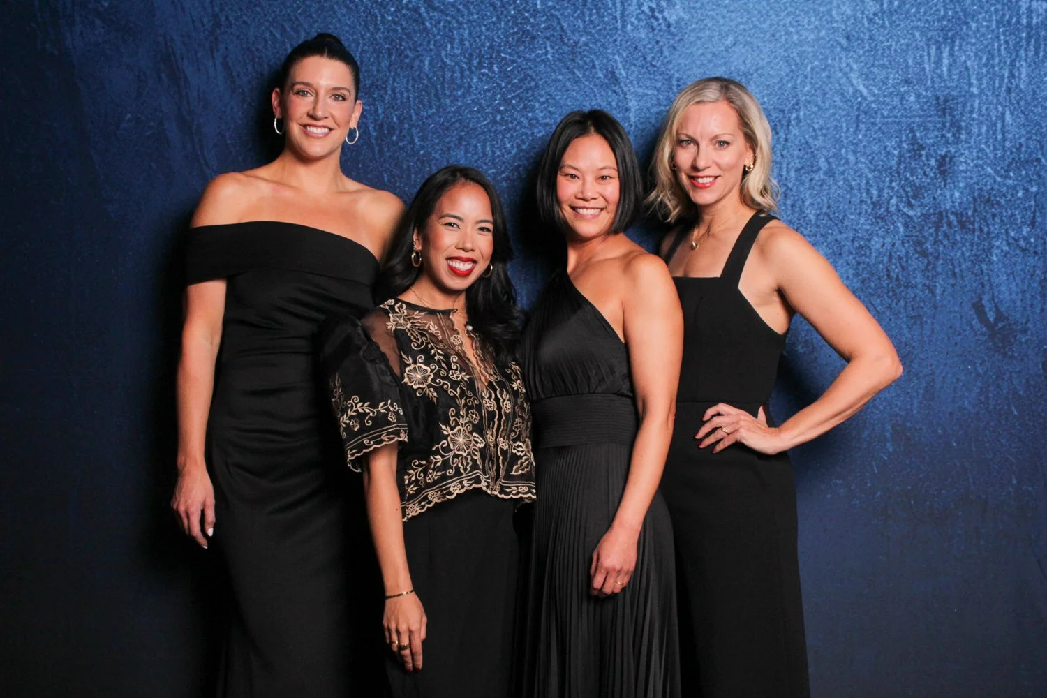 Four women dressed in elegant black dresses posing together against a dark blue background.