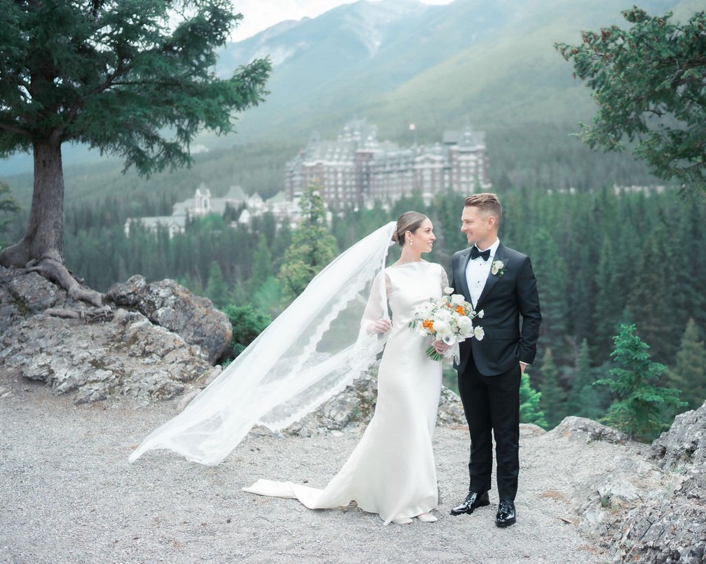 A bride and groom standing outdoors on rocky terrain with a forested mountain landscape and a large castle in the background, posing for a wedding photo.