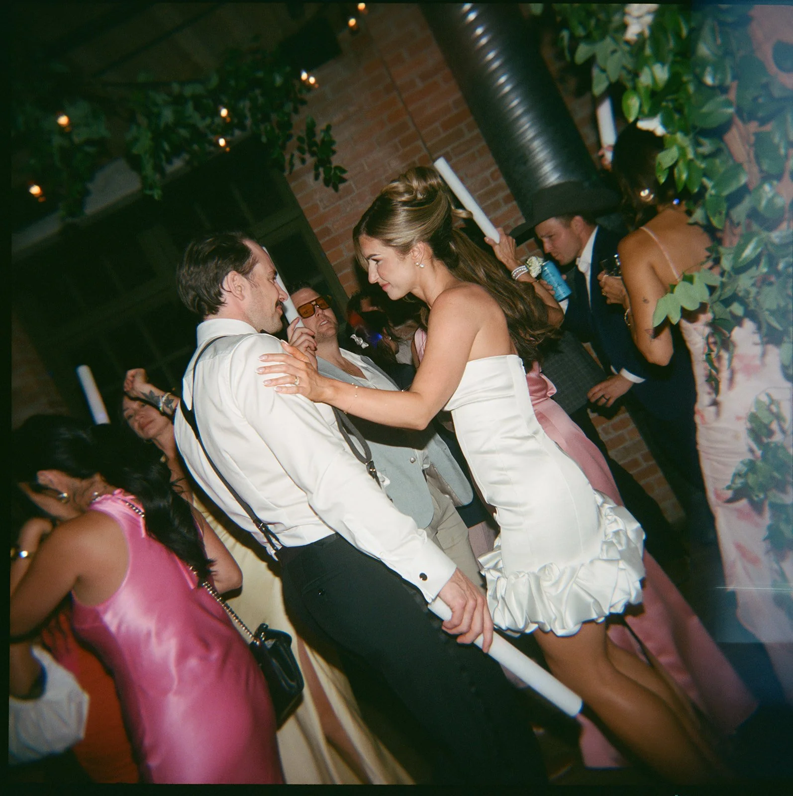 People dancing at a party with a brick wall background, including a woman in a white dress and a man in a white shirt, surrounded by others in formal attire.