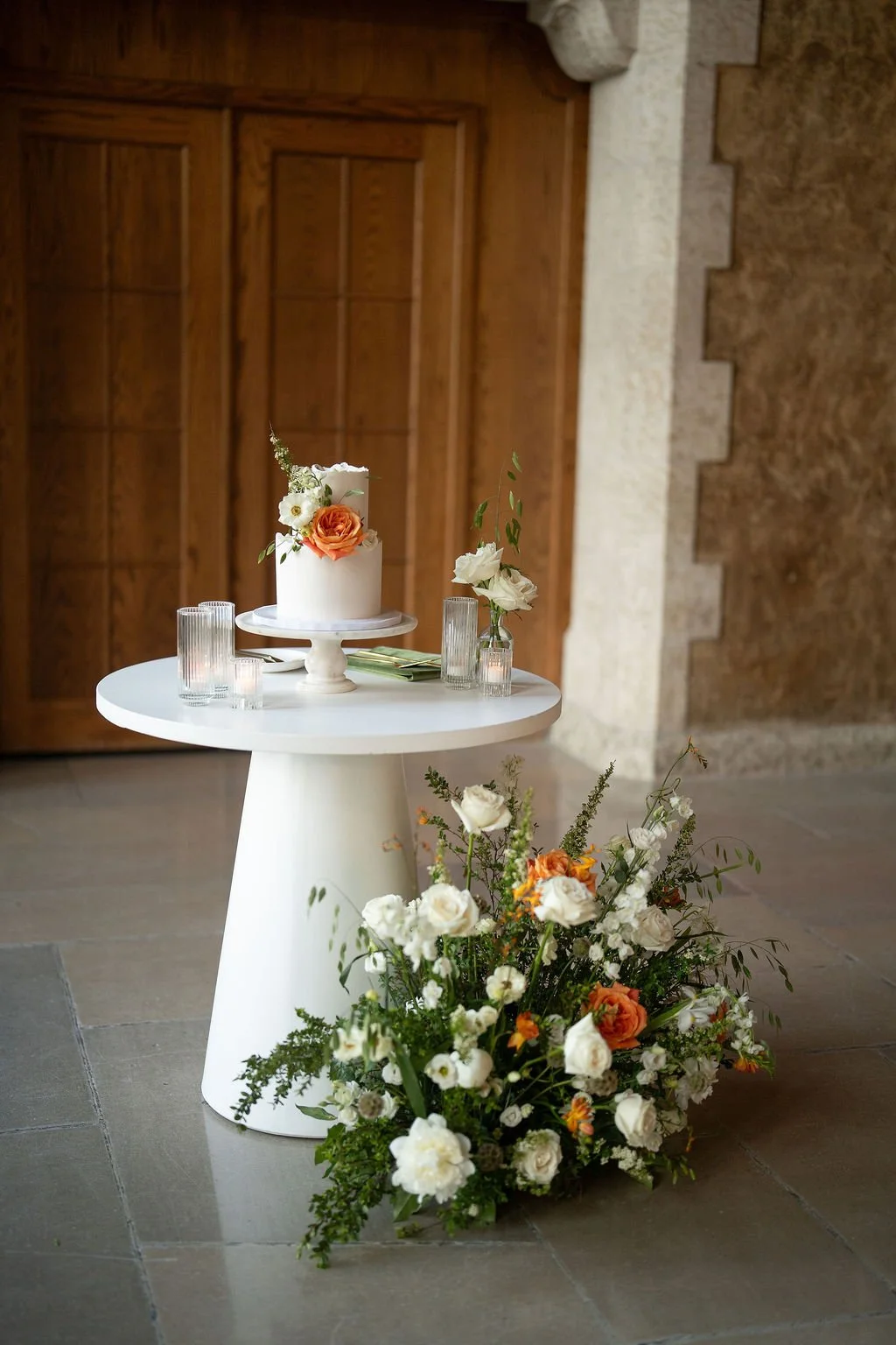 A white cake decorated with flowers on a white cake stand, sitting on a round white table. Next to it are glassware and a small floral arrangement in a glass vase. A large floral bouquet is placed on the floor beside the table, featuring white and or